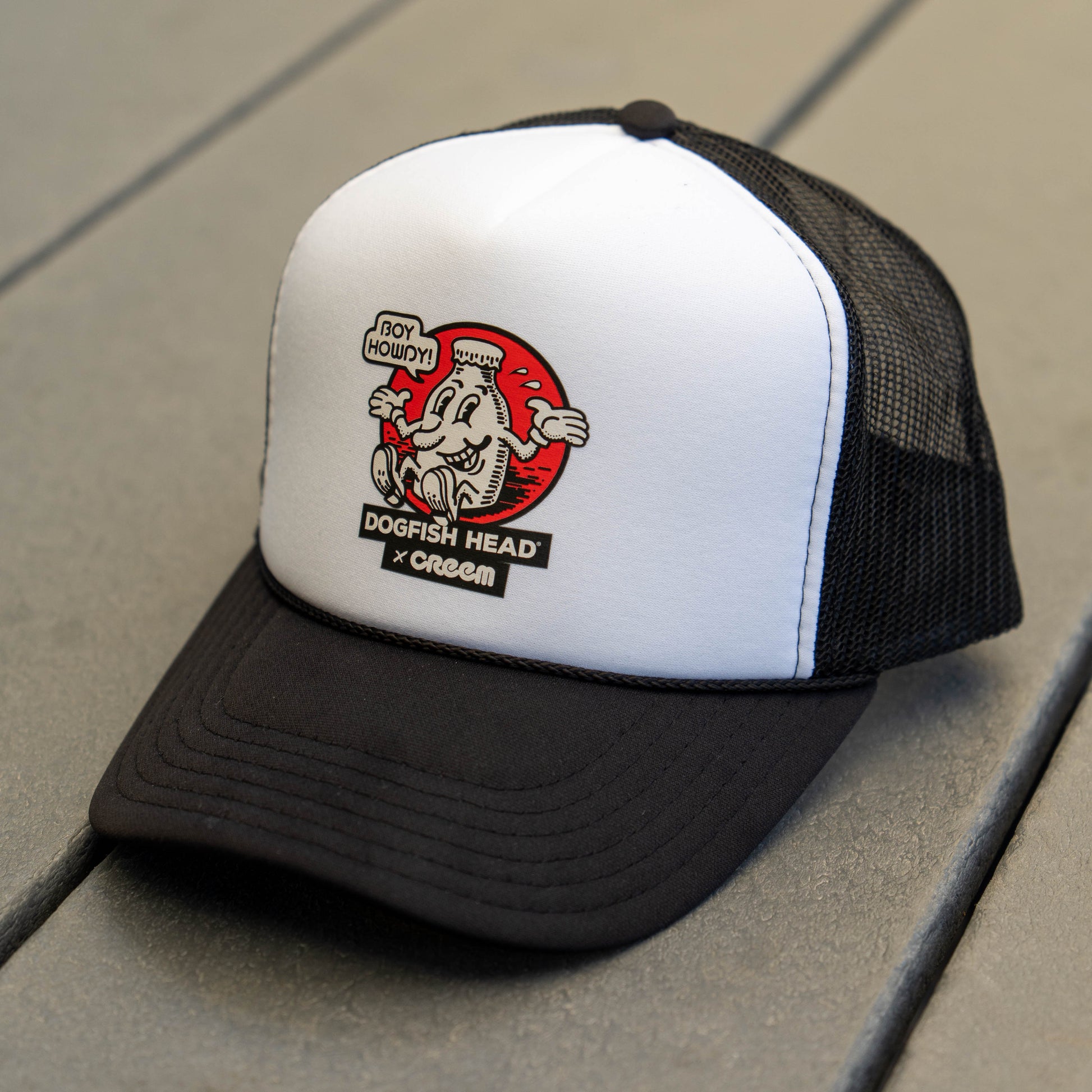 A black and white trucker hat with a mesh back, resting on a gray wooden surface. The front panel is white with a black brim and features a graphic with the text 'DOGFISH HEAD x Creem' and 'BOY HOWDY'.