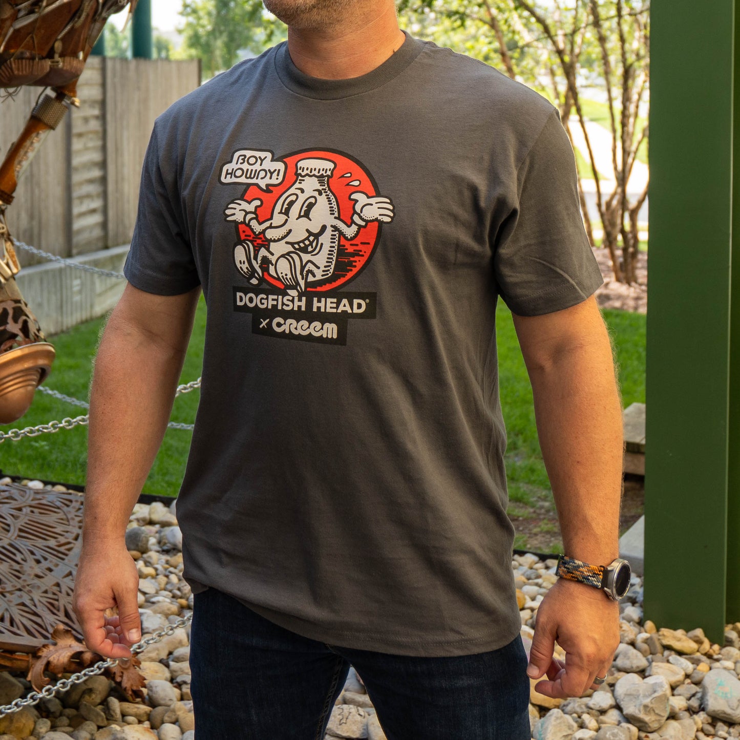 A man wearing a dark gray graphic t-shirt featuring a cartoon dogfish head with the text 'BOY HOWDY' and 'DOGFISH HEAD x Creem'. The photo is taken outdoors in a backyard setting with a wooden fence and greenery in the background.