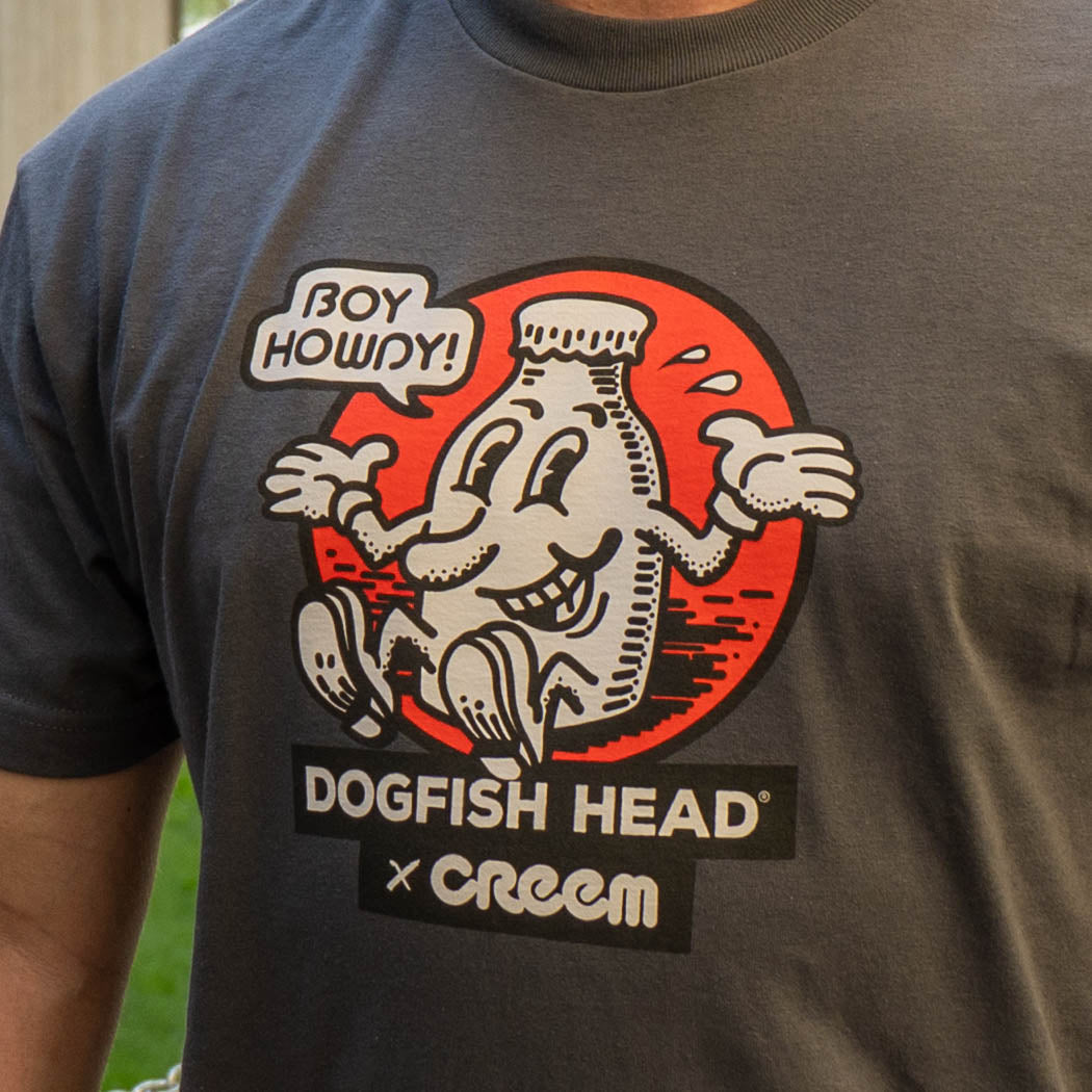 A close-up of a person wearing a dark gray t-shirt featuring a graphic print of the Dogfish Head x Creem collaboration. The graphic shows a white graphic inside a red circle with a speech bubble that reads 'BOY HOWDY!'.