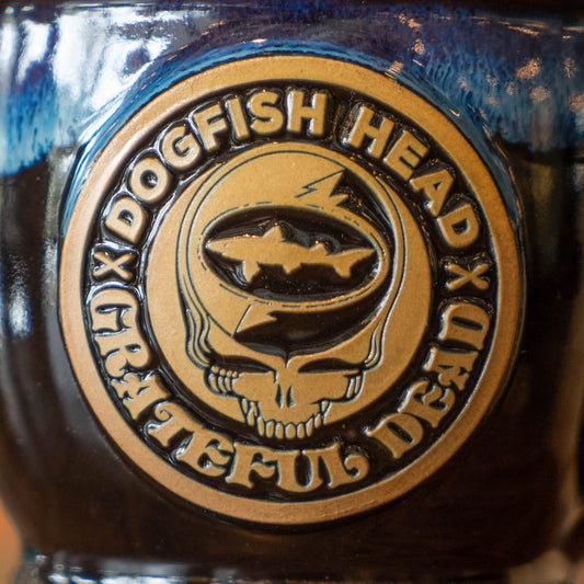 Close up of logo on ceramic mug. Logo is brown with "Dogfish Head x Grateful Dead" in a circle. In the circle is a Grateful Dead skull with a Dogfish head logo between the lightning bolts. 