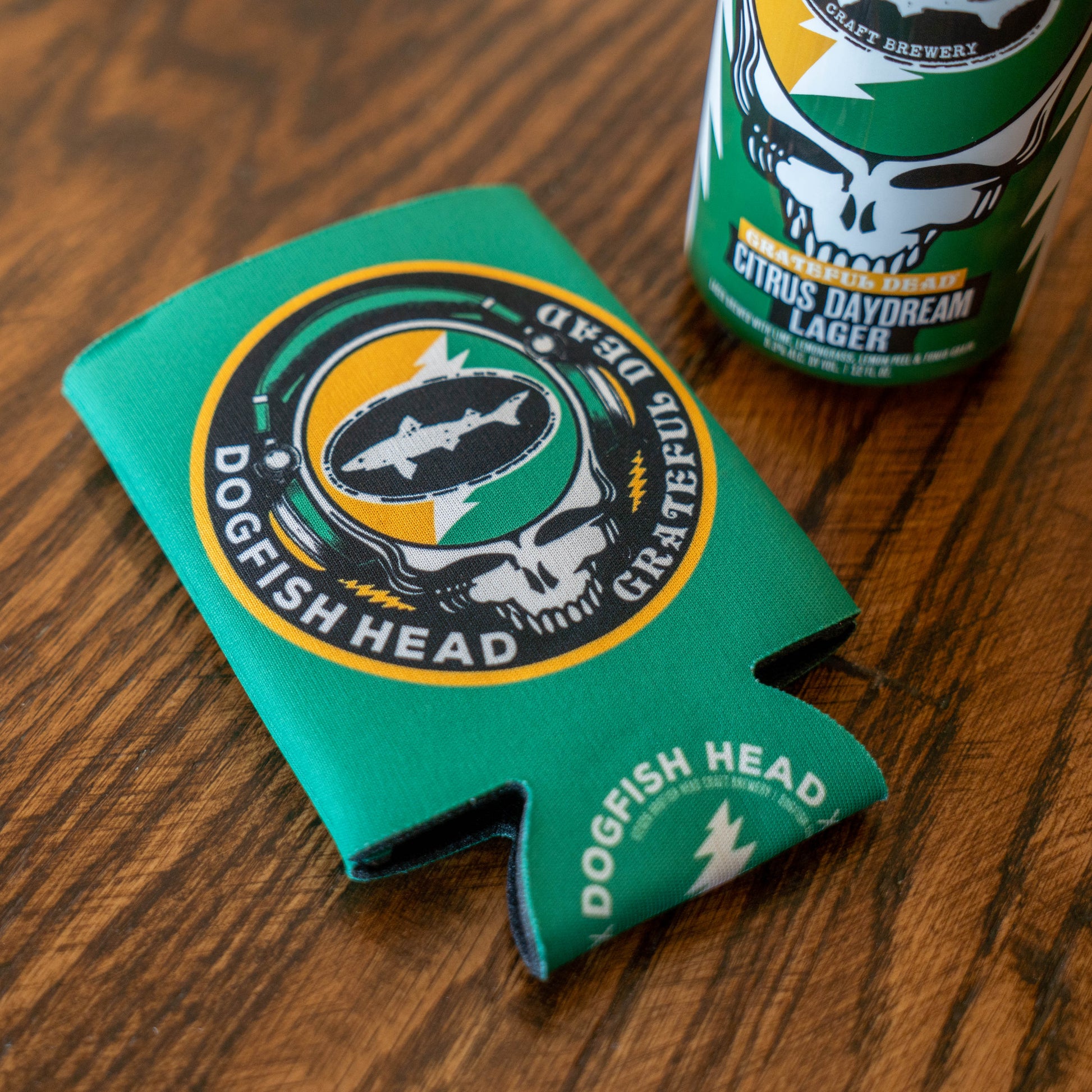 A Dogfish Head x Grateful Dead Citrus Daydream 12oz Can Cooler by Grandstand Glassware & Apparel sits beside a matching lager can, both featuring coordinating artwork and Grateful Dead logos on a wooden surface.