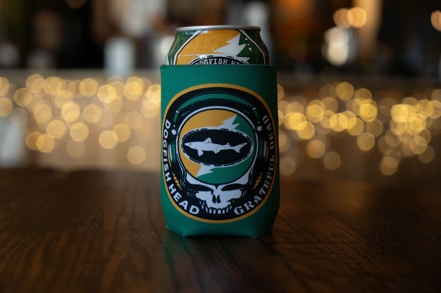 A Dogfish Head x Grateful Dead Citrus Daydream 12oz Can Cooler by Grandstand Glassware & Apparel, featuring a fish, skull, and lightning bolt logo, sits on a wooden table with blurred lights in the background.