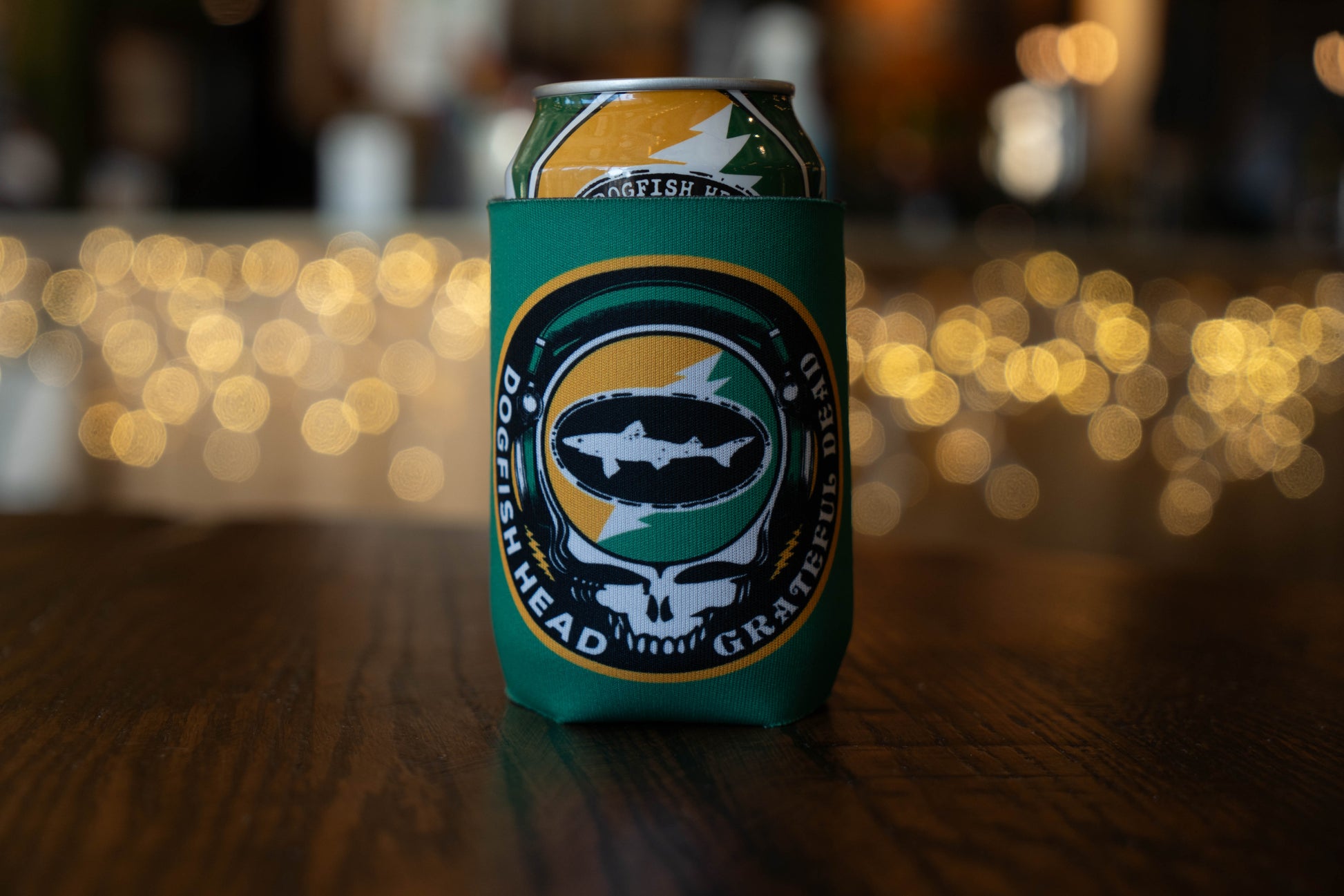 A Dogfish Head x Grateful Dead Citrus Daydream 12oz Can Cooler by Grandstand Glassware & Apparel, featuring a fish, skull, and lightning bolt logo, sits on a wooden table with blurred lights in the background.