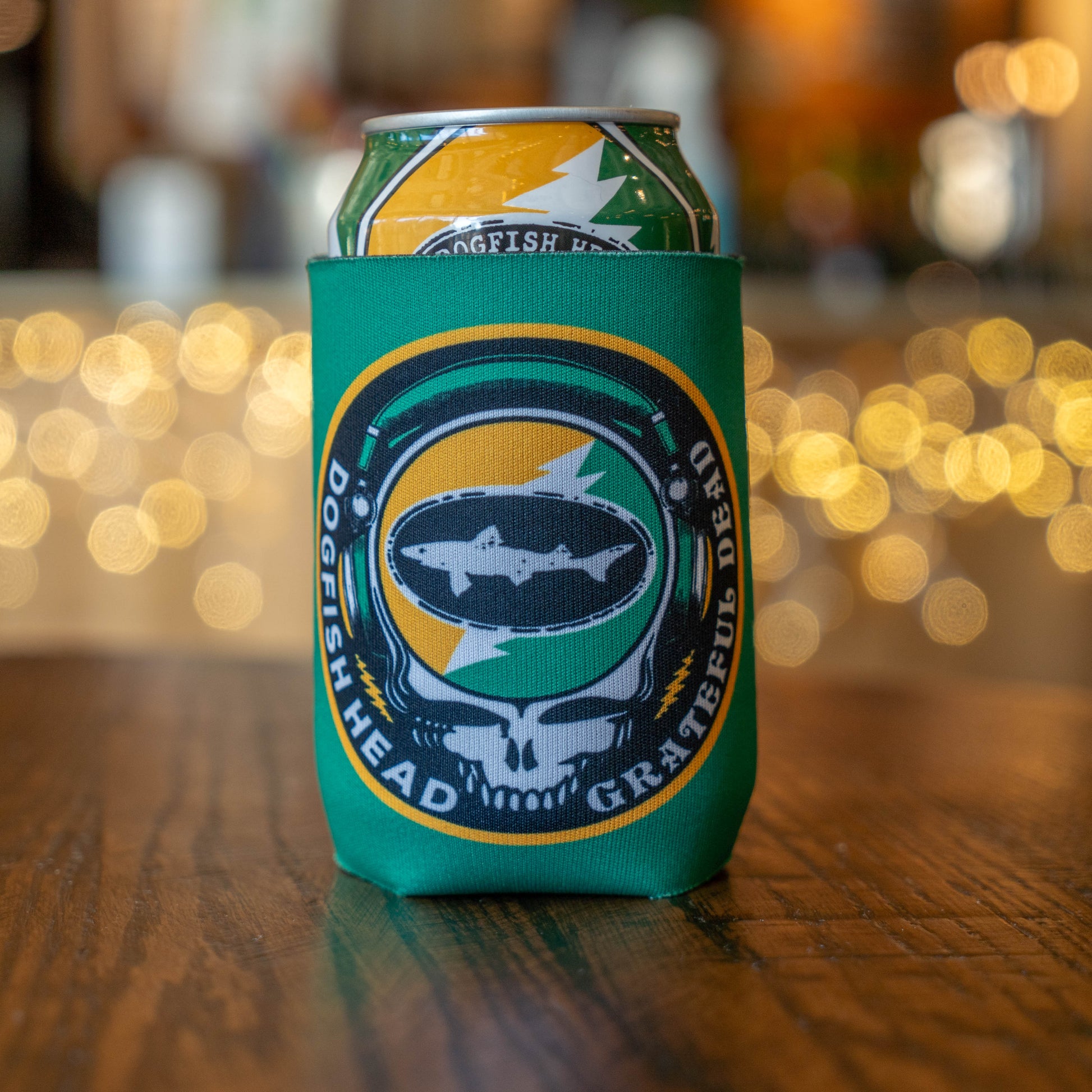 A Dogfish Head x Grateful Dead Citrus Daydream 12oz Can Cooler by Grandstand Glassware & Apparel, featuring a skull, fish, and lightning bolt design, holds a beer can on a wooden table with blurred lights in the background.