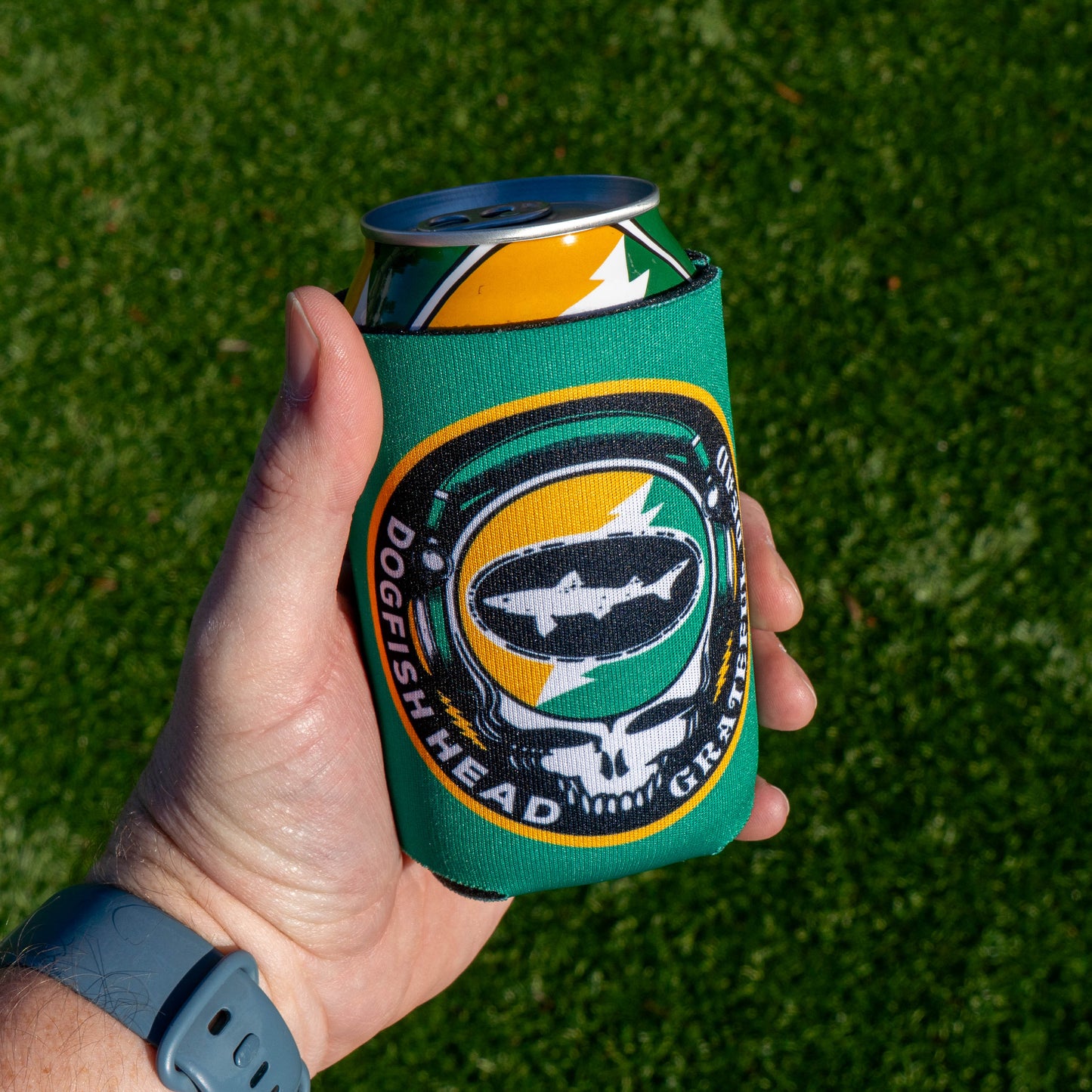 A hand wearing a gray watch holds a can in the Dogfish Head x Grateful Dead Citrus Daydream 12oz Can Cooler by Grandstand Glassware & Apparel, with green grass in the background.