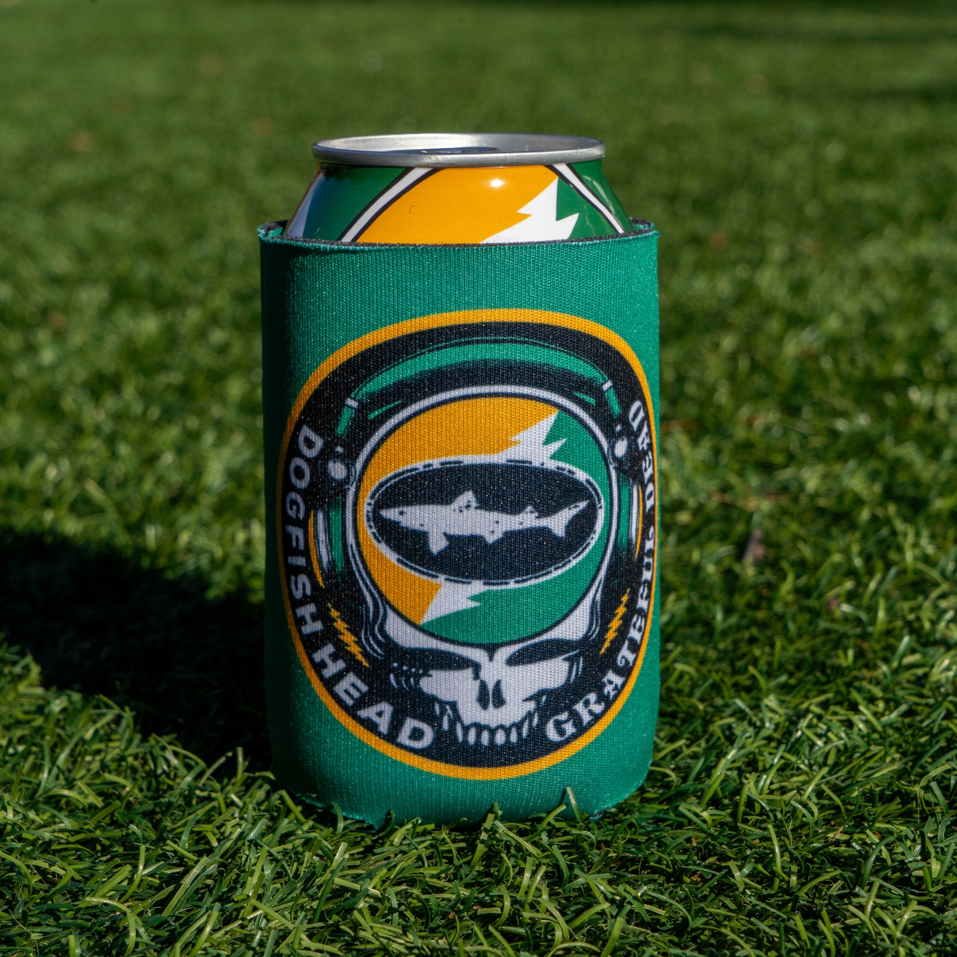 A Dogfish Head x Grateful Dead Citrus Daydream 12oz Can Cooler by Grandstand Glassware & Apparel sits on grass, its colorful skull and fish design catching sunlight as it shades a can.