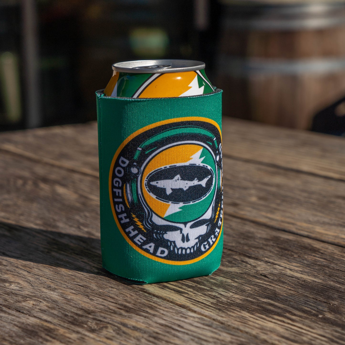 A Dogfish Head x Grateful Dead Citrus Daydream 12oz Can Cooler by Grandstand Glassware & Apparel sits on a wooden table in sunlight, with blurred barrels and other objects in the background.