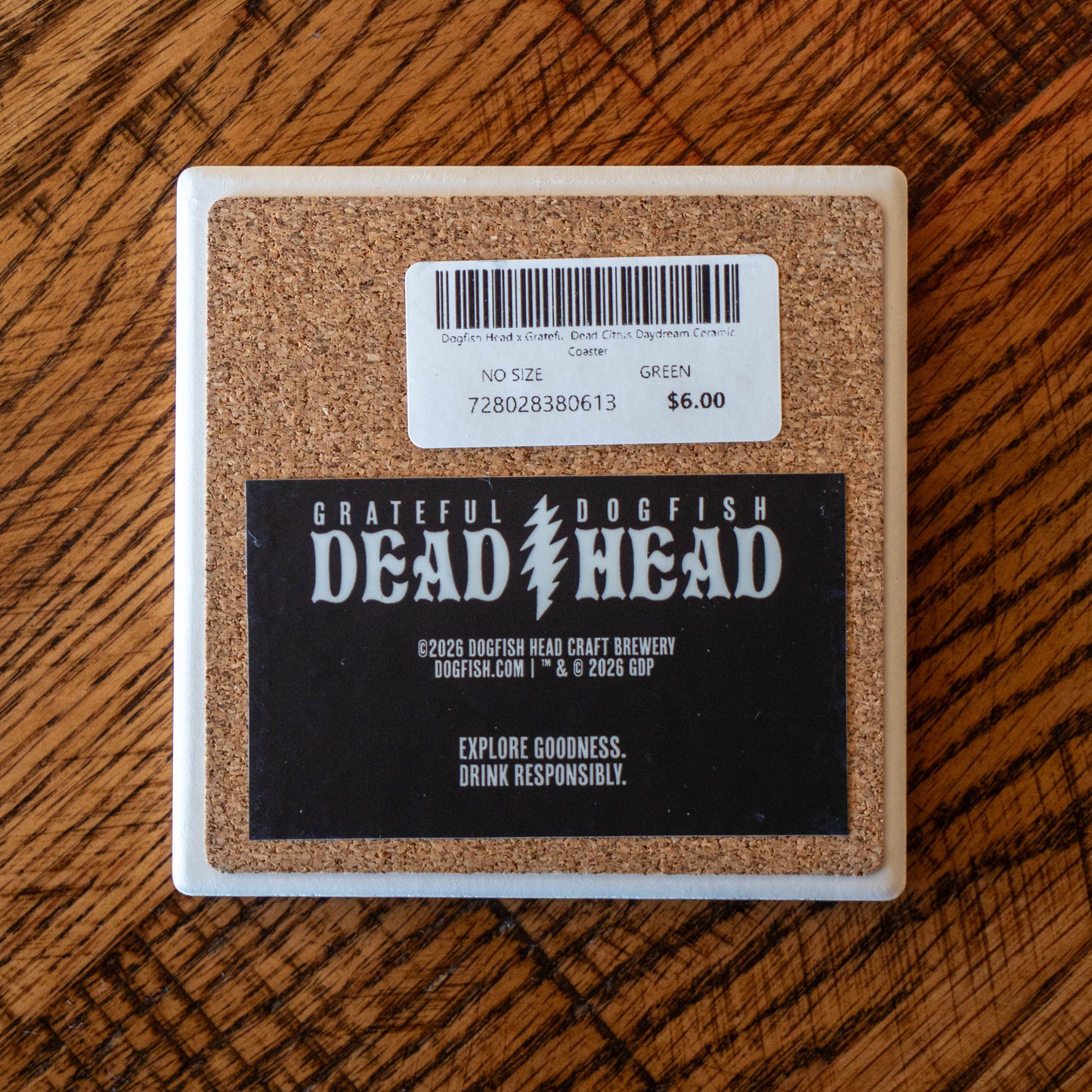 The Dogfish Head x Grateful Dead Citrus Daydream Ceramic Coaster by Paint the Town, Inc. lies on wood, displaying a “GRATEFUL DOGFISH DEAD HEAD” label, “Explore Goodness. Drink Responsibly.” message, $6 price tag, and barcode.