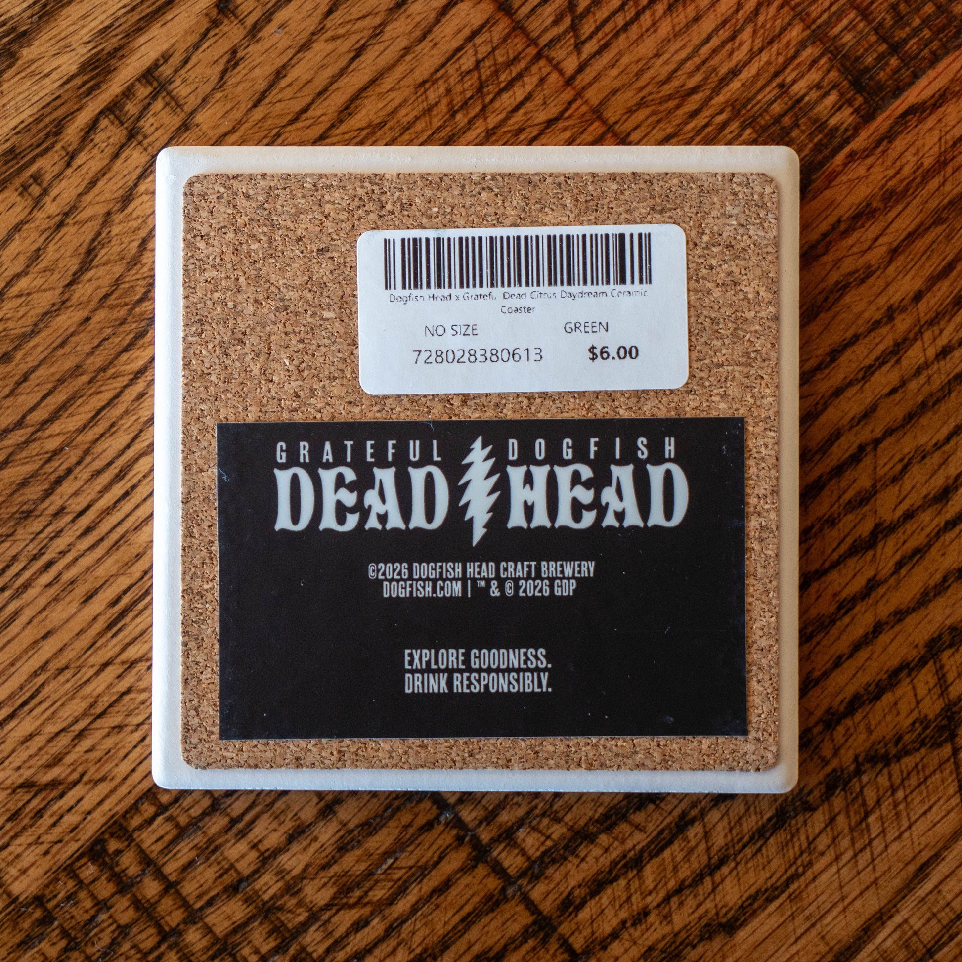 The Dogfish Head x Grateful Dead Citrus Daydream Ceramic Coaster by Paint the Town, Inc. lies on wood, displaying a “GRATEFUL DOGFISH DEAD HEAD” label, “Explore Goodness. Drink Responsibly.” message, $6 price tag, and barcode.