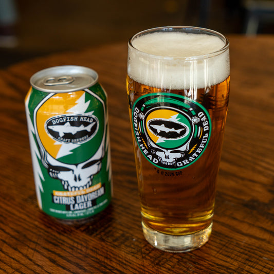 A Dogfish Head x Grateful Dead Citrus Daydream Glass by Grandstand Glassware & Apparel, filled with golden beer and topped with a foamy head, sits beside a matching can on a wooden table.