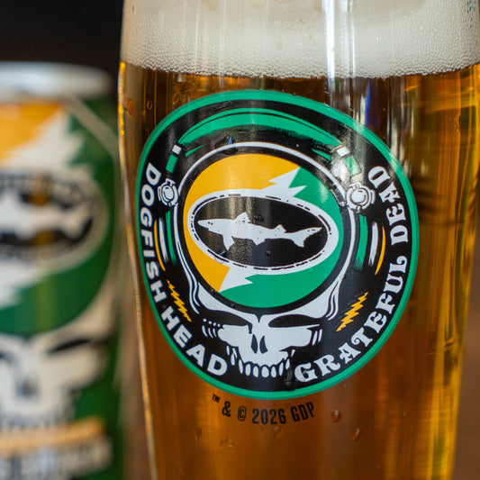 A close-up of the Dogfish Head x Grateful Dead Citrus Daydream Glass by Grandstand Glassware & Apparel, filled with amber beer and featuring a skull and lightning bolt logo; a matching can appears blurred in the background.