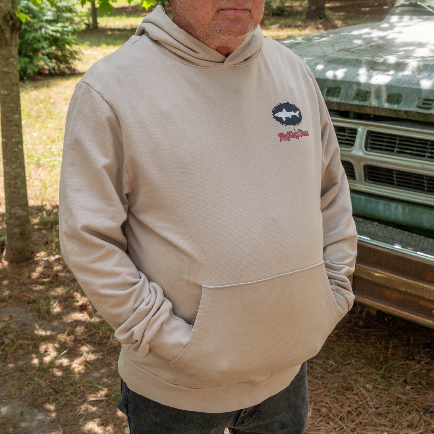 A person wearing the Dogfish Head x Rolling Stone Mocha Hoodie by GO WEST T-SHIRT COMPANY with a front pocket and graphic designs stands outdoors near an old vehicle, with trees and grass in the background. Their face is partially out of frame.