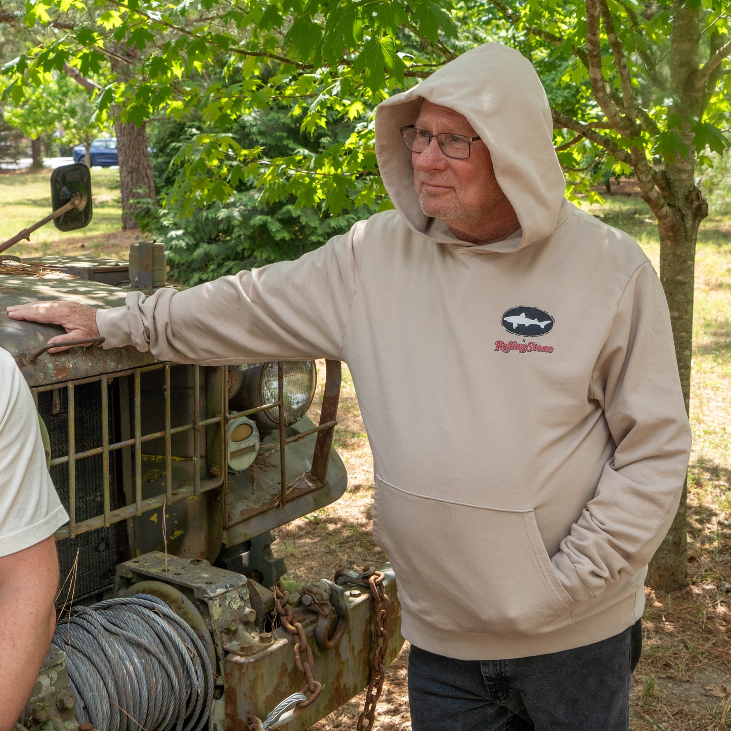An older man in a Dogfish Head x Rolling Stone Mocha Hoodie by GO WEST T-SHIRT COMPANY leans against a vintage off-road vehicle in a sunlit, wooded setting.