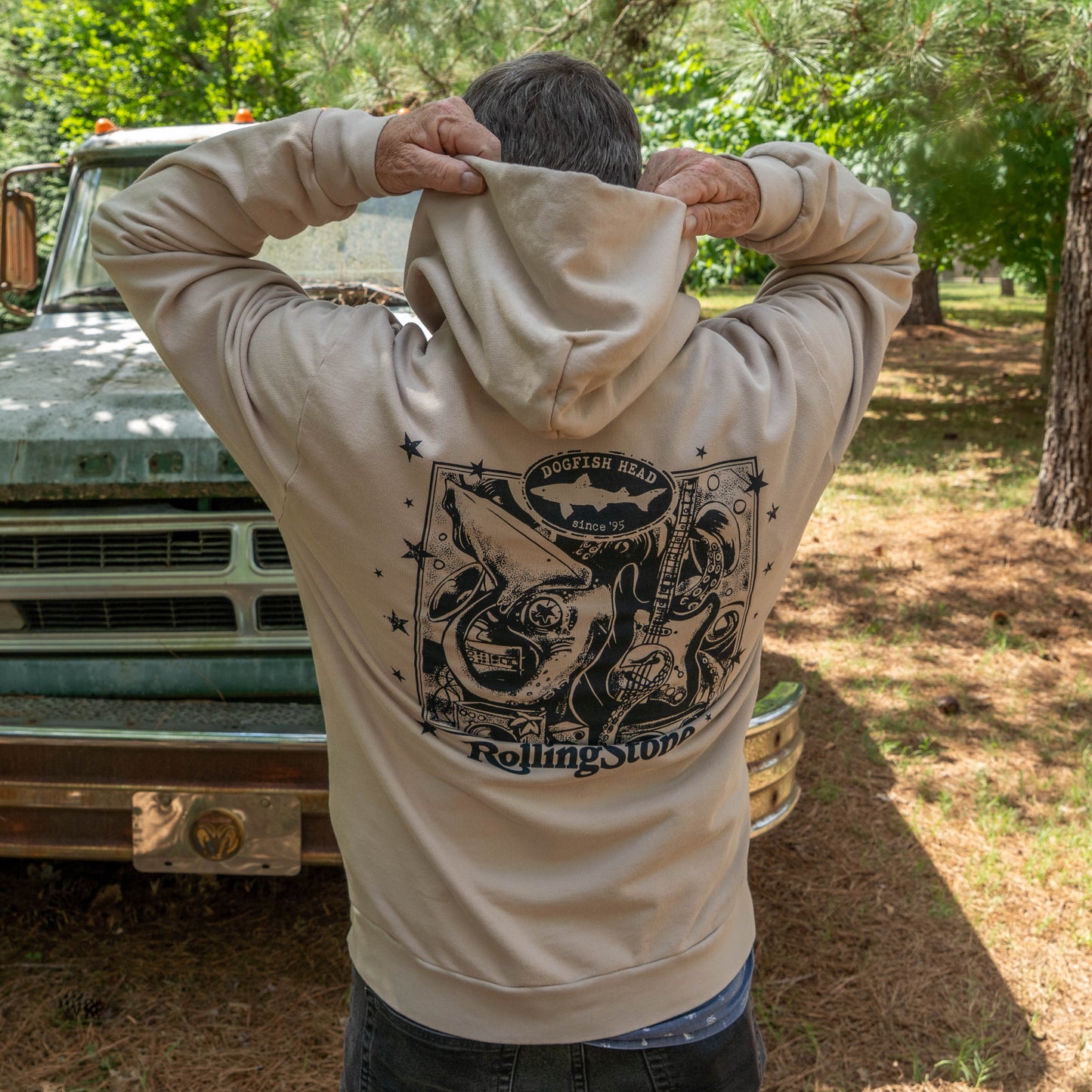 A person outdoors by an old green truck lifts the hood of a tan Dogfish Head x Rolling Stone Mocha Hoodie by GO WEST T-SHIRT COMPANY, featuring a bold black graphic and logo on the back. Trees and grass are in the background.