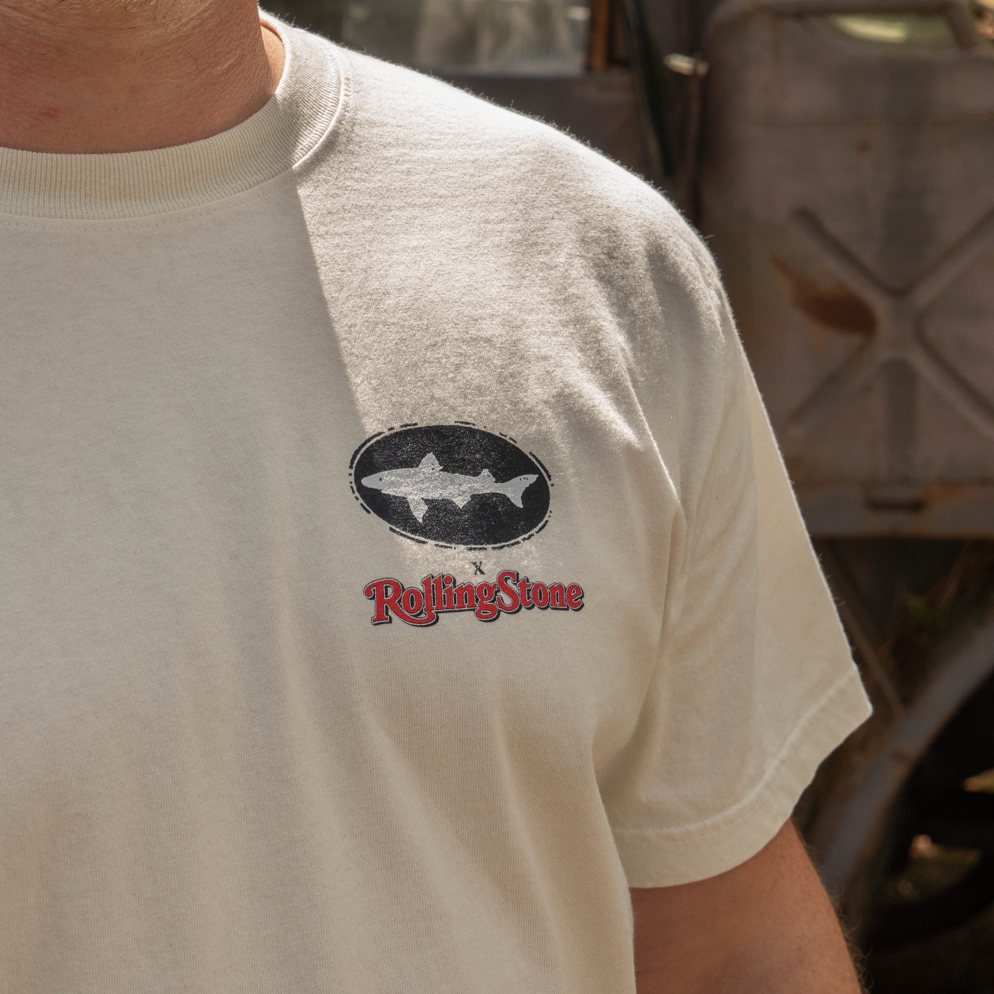 A person wearing the Dogfish Head x Rolling Stone Oatmeal Tee Shirt by GO WEST T-SHIRT COMPANY, featuring a black oval shark logo above red Rolling Stone text, stands outdoors near a rusty vehicle. Only the upper body is visible.