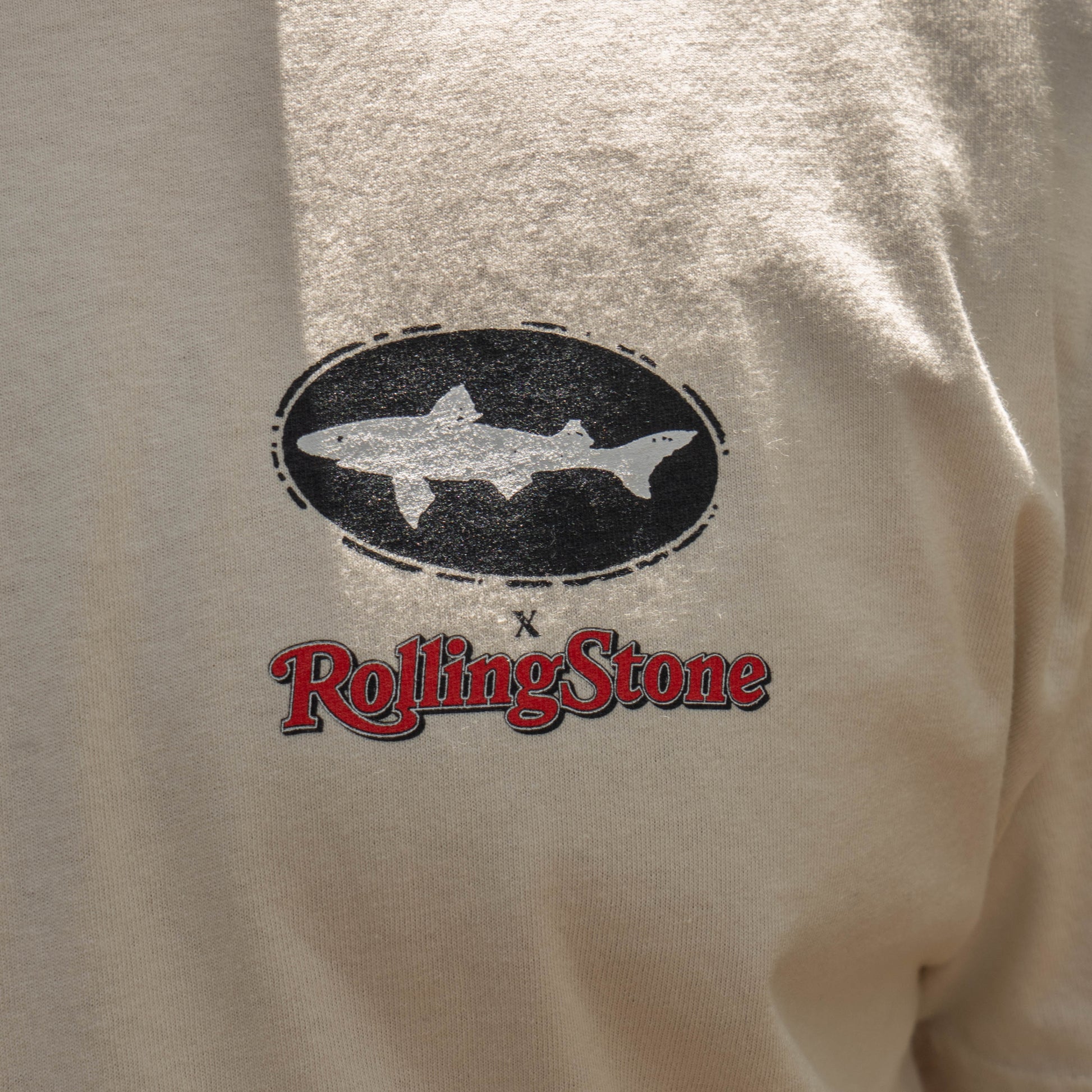 The Dogfish Head x Rolling Stone Oatmeal Tee Shirt by GO WEST T-SHIRT COMPANY features a black oval with a white shark silhouette above the red Rolling Stone logo on a beige background.