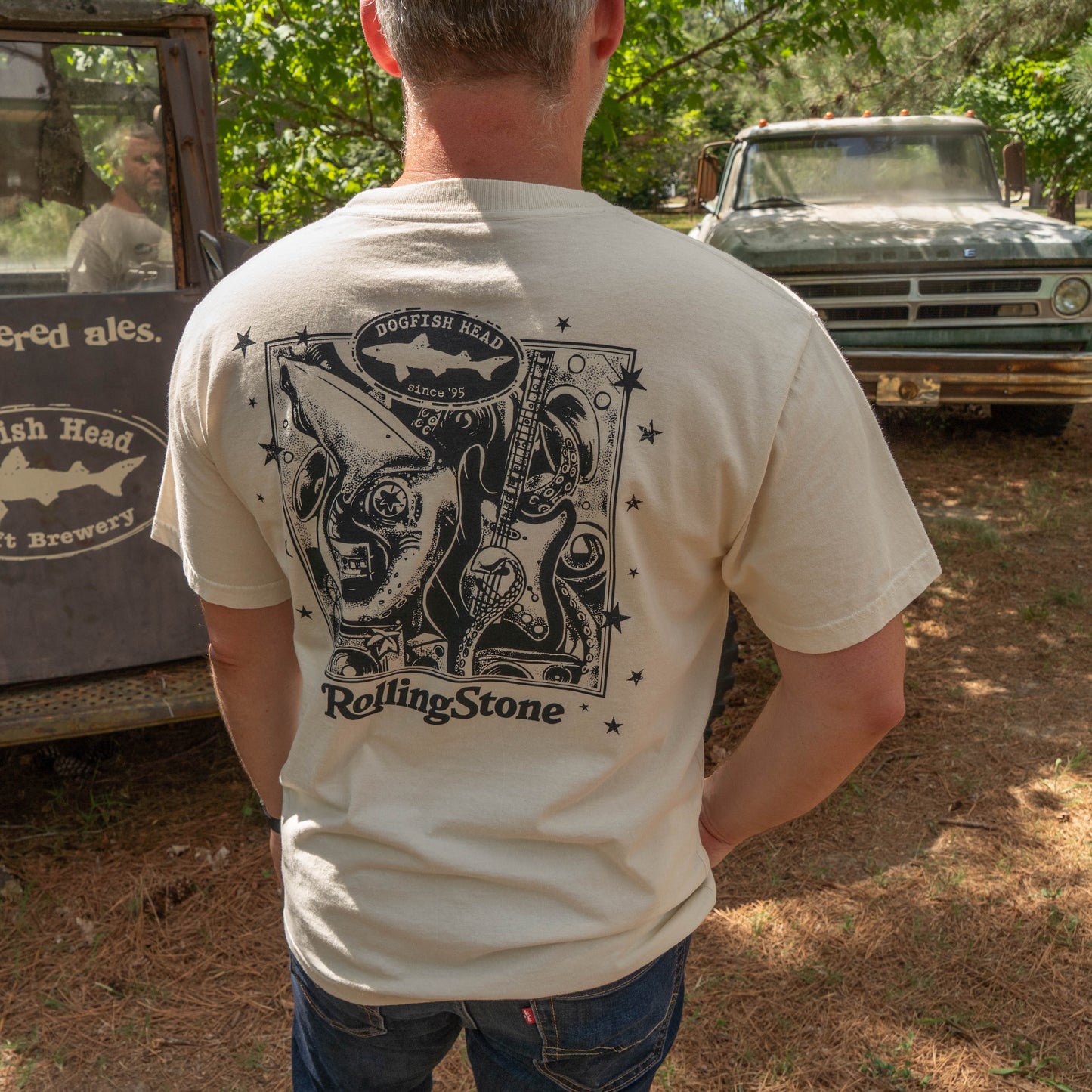 A man outdoors, back turned, wears a GO WEST T-SHIRT COMPANY Dogfish Head x Rolling Stone Oatmeal Tee Shirt. Two vintage trucks are parked nearby in a sunny wooded area.