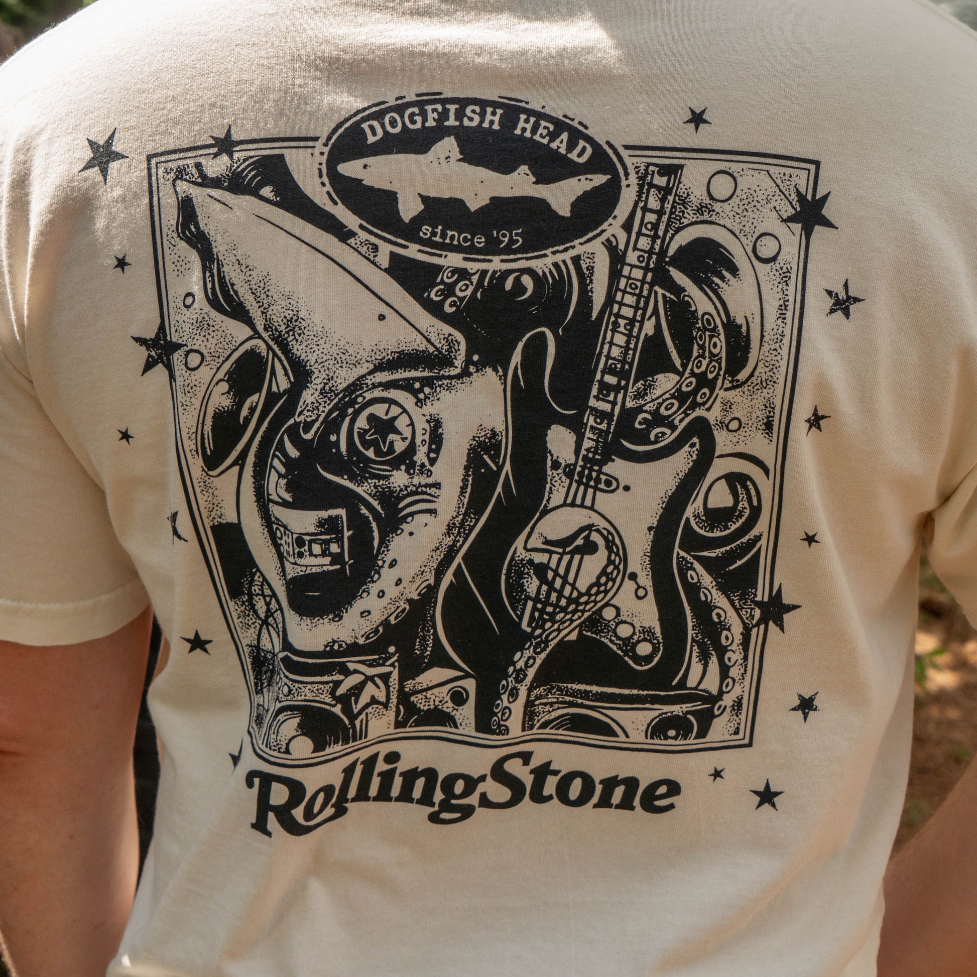The back of the cream-colored Dogfish Head x Rolling Stone Oatmeal Tee Shirt by GO WEST T-SHIRT COMPANY features a black graphic with a shark, guitar, stars, and "Dogfish Head since 95" and "Rolling Stone.