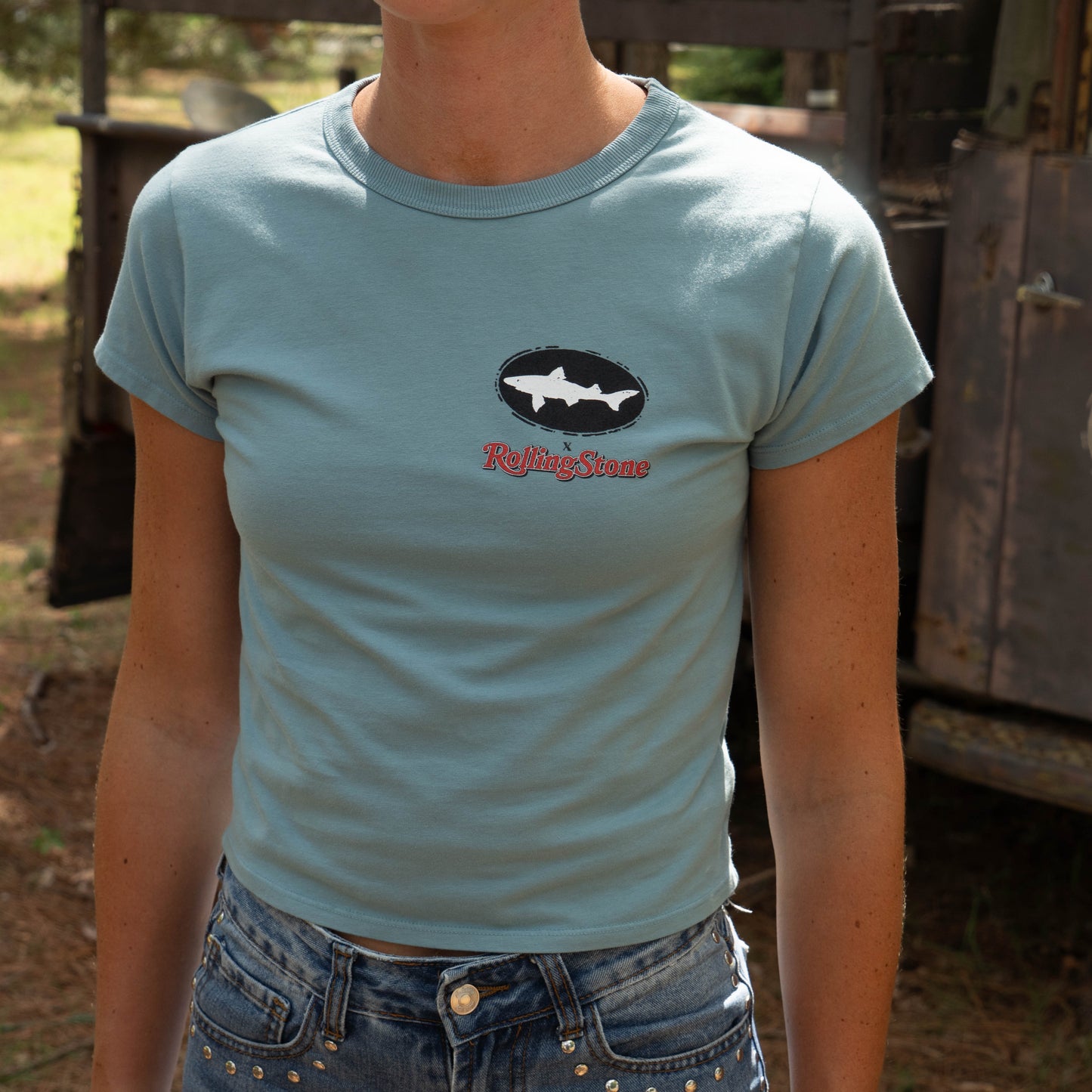 Outdoors, someone wears the Dogfish Head x Rolling Stone Women's Slate Blue Baby Tee Shirt by GO WEST T-SHIRT COMPANY with blue jeans featuring silver studs. Blurred greenery and a rustic vehicle appear in the background.