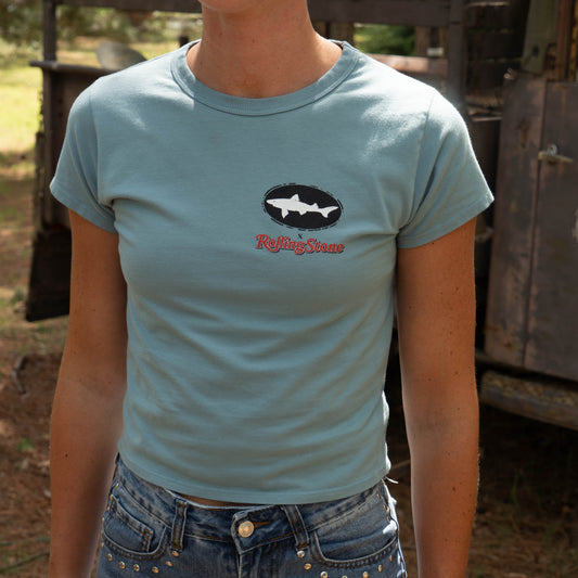 A person in a Dogfish Head x Rolling Stone Slate Blue Baby Tee Shirt by GO WEST T-SHIRT COMPANY stands outdoors beside a rustic vehicle, with their face not visible.