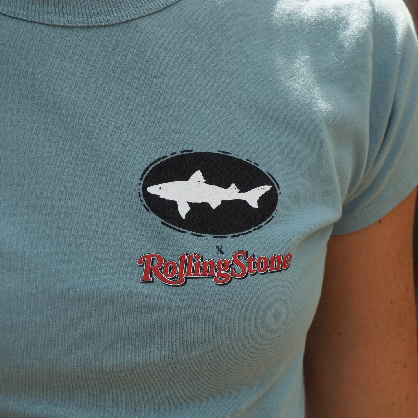 The Dogfish Head x Rolling Stone Women's Slate Blue Baby Tee Shirt by GO WEST T-SHIRT COMPANY features a white shark graphic above the red Rolling Stone logo on the chest, creating a classic rock and roll look.