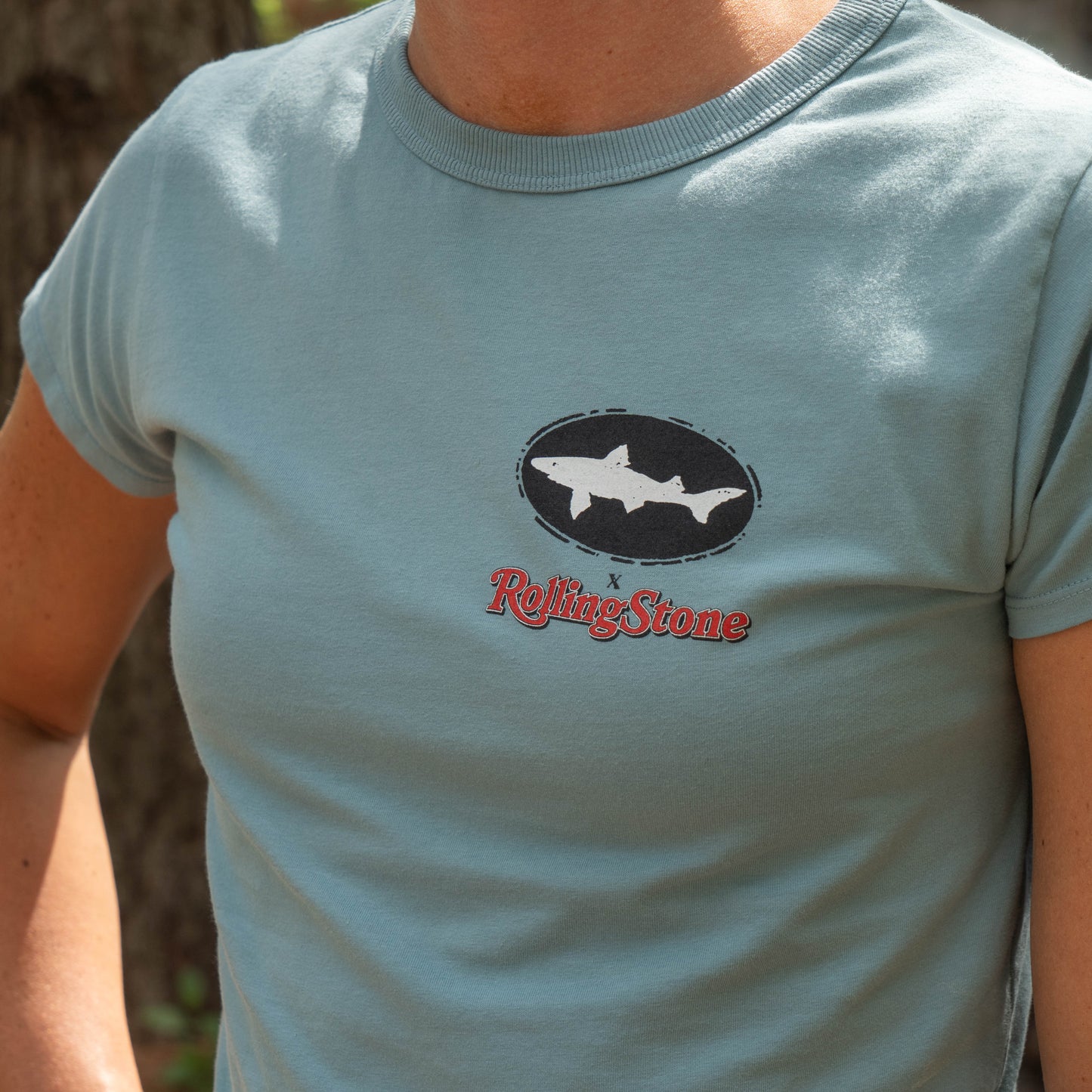 A person wearing a GO WEST T-SHIRT COMPANY Dogfish Head x Rolling Stone Women's Slate Blue Baby Tee Shirt with a shark graphic above the red Rolling Stone logo. Only the torso and part of the arms are visible.