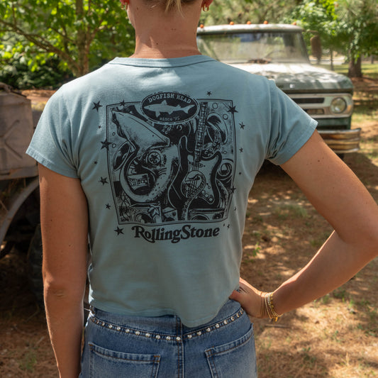 A person wearing a Dogfish Head x Rolling Stone Slate Blue Baby Tee Shirt by GO WEST T-SHIRT COMPANY stands outdoors with their back to the camera. Old trucks and trees are seen in the background.