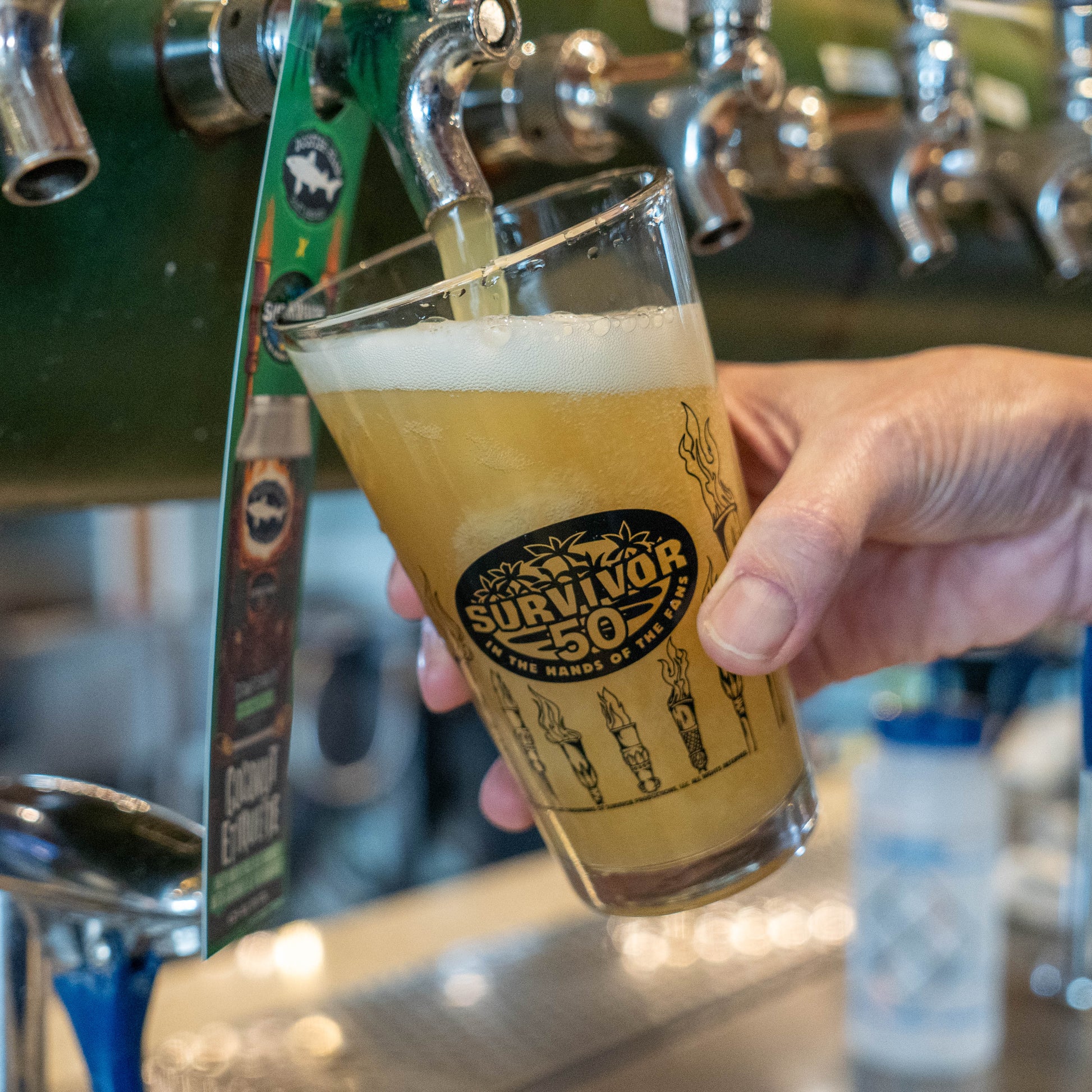 Person holding a glass of beer with 'Survivor 50' label at a bar.