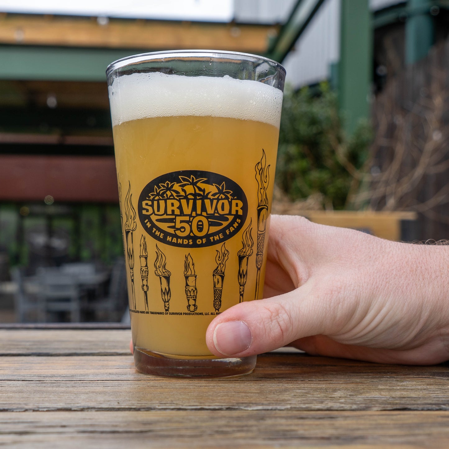 Hand holding a glass of beer with 'Survivor 50' logo outdoors