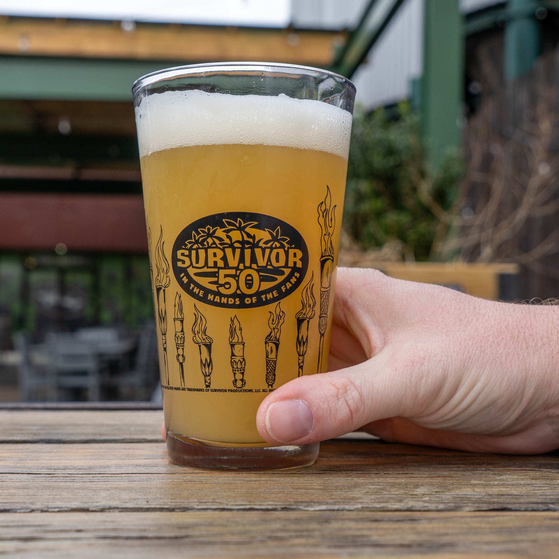 Hand holding a glass of beer with 'Survivor 50' logo outdoors