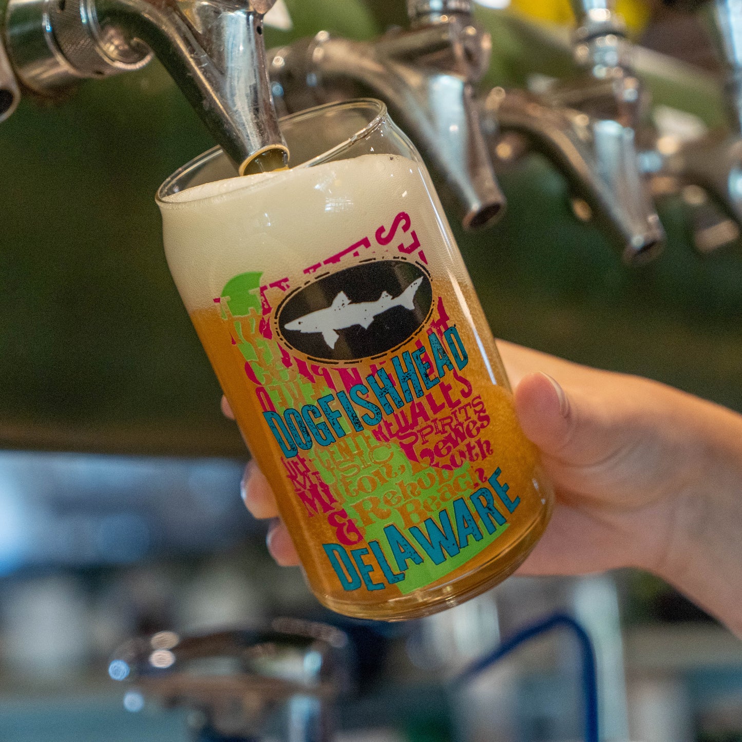 A hand holds a clear pint glass with a colorful 'Dogfish Head Delaware' label under a metal beer tap, with beer foaming into the glass in a bar setting.