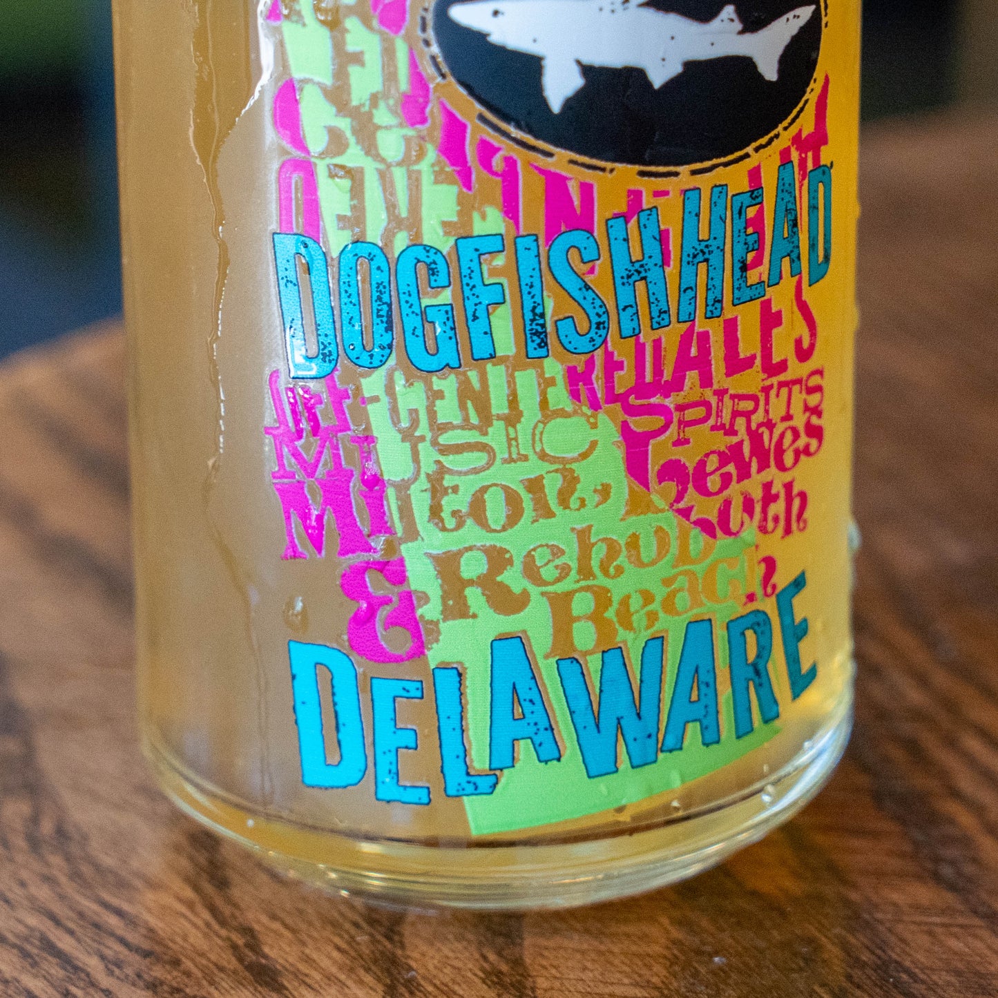 A close-up of a clear glass bottle or tumbler with a colorful printed label featuring the words 'Dogfish Head' and 'Delaware' in bright blue and pink lettering, placed on a wooden surface.