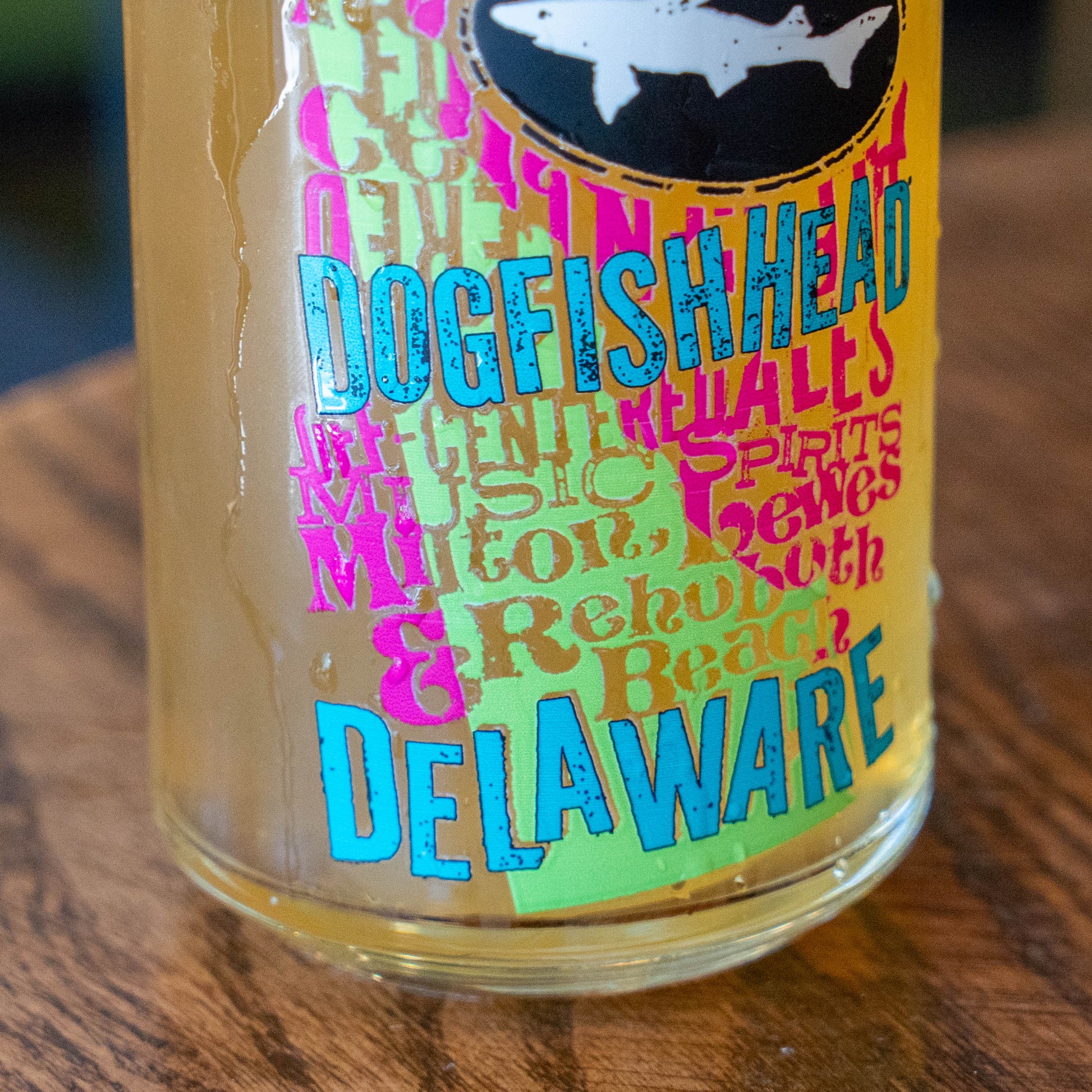 A close-up of a clear glass bottle or tumbler with a colorful printed label featuring the words 'Dogfish Head' and 'Delaware' in bright blue and pink lettering, placed on a wooden surface.
