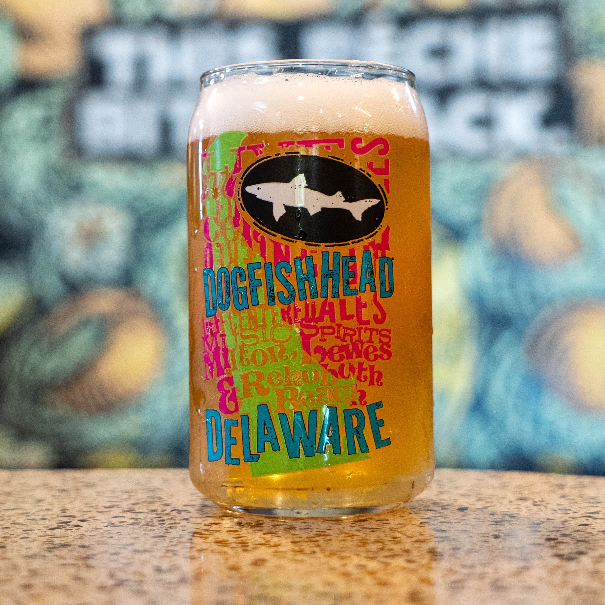 A clear glass can-shaped beer mug filled with golden beer, featuring a colorful Dogfish Head Delaware label with a shark logo, set on a speckled countertop with a blurred background.