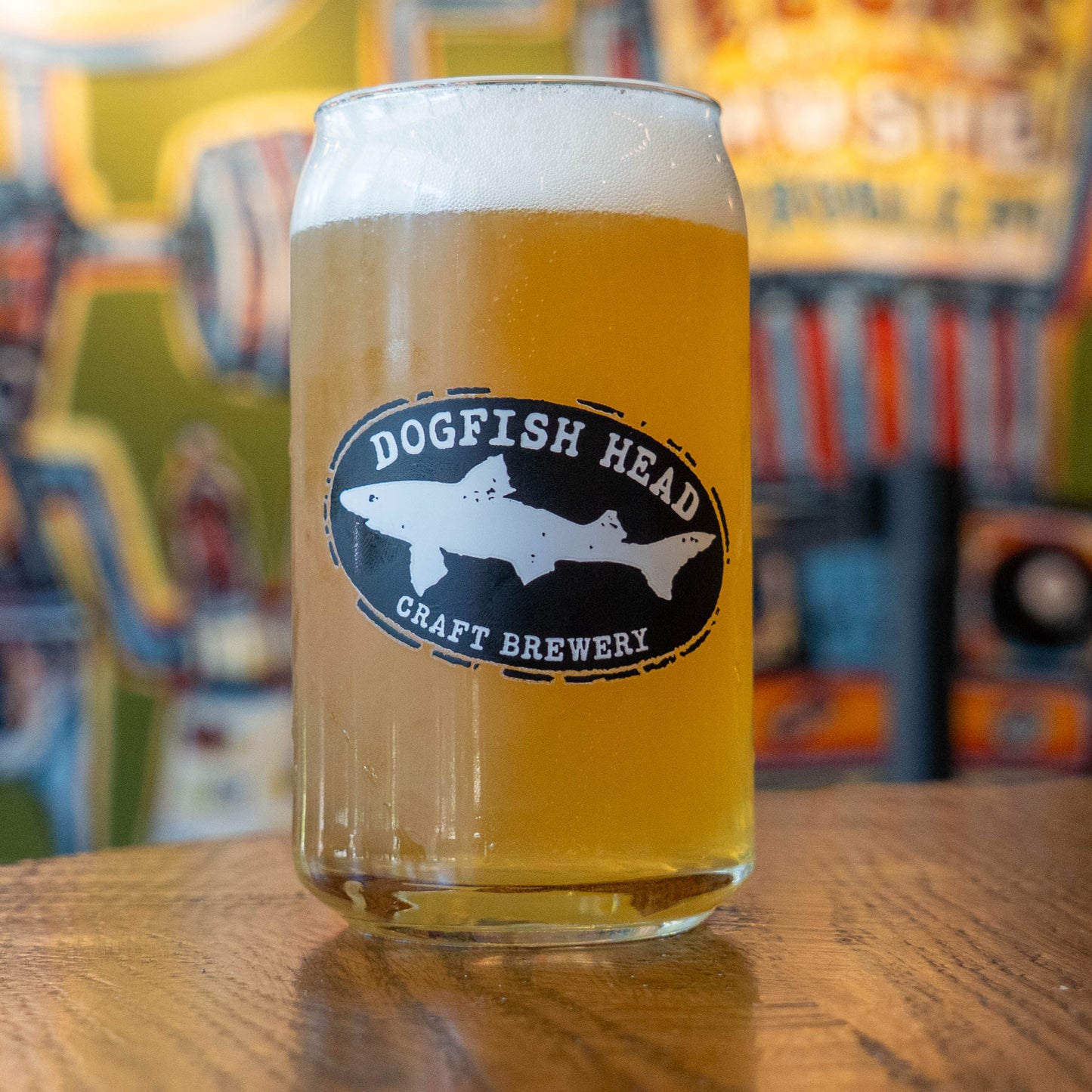 A clear glass pint mug filled with golden beer and topped with white foam, featuring a black oval Dogfish Head Craft Brewery logo on the front, set on a wooden table with a blurred bar background.