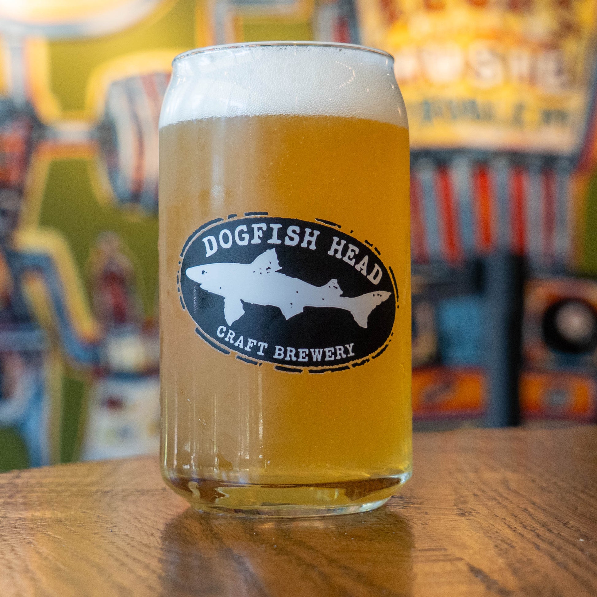 A clear glass pint mug filled with golden beer and topped with white foam, featuring a black oval Dogfish Head Craft Brewery logo on the front, set on a wooden table with a blurred bar background.
