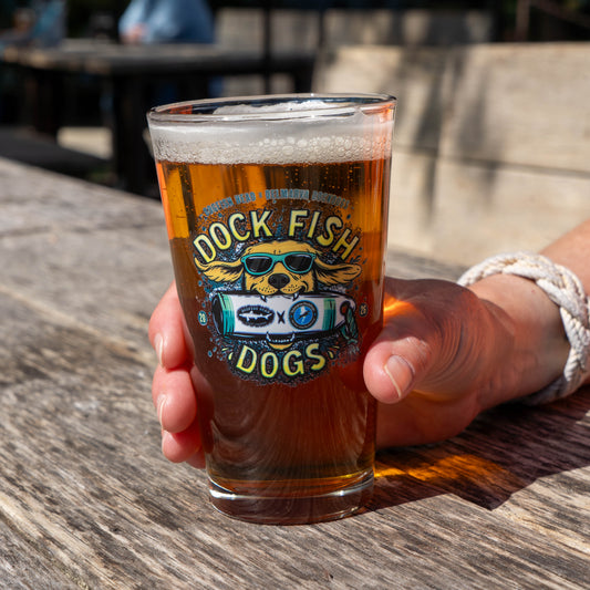 Man holding pint glass with a graphic of a golden retriever wearing sunglasses biting into Dogfish Head x Delmarva Dock Dogs plushie. "Dog Fish Dogs" written in yellow 