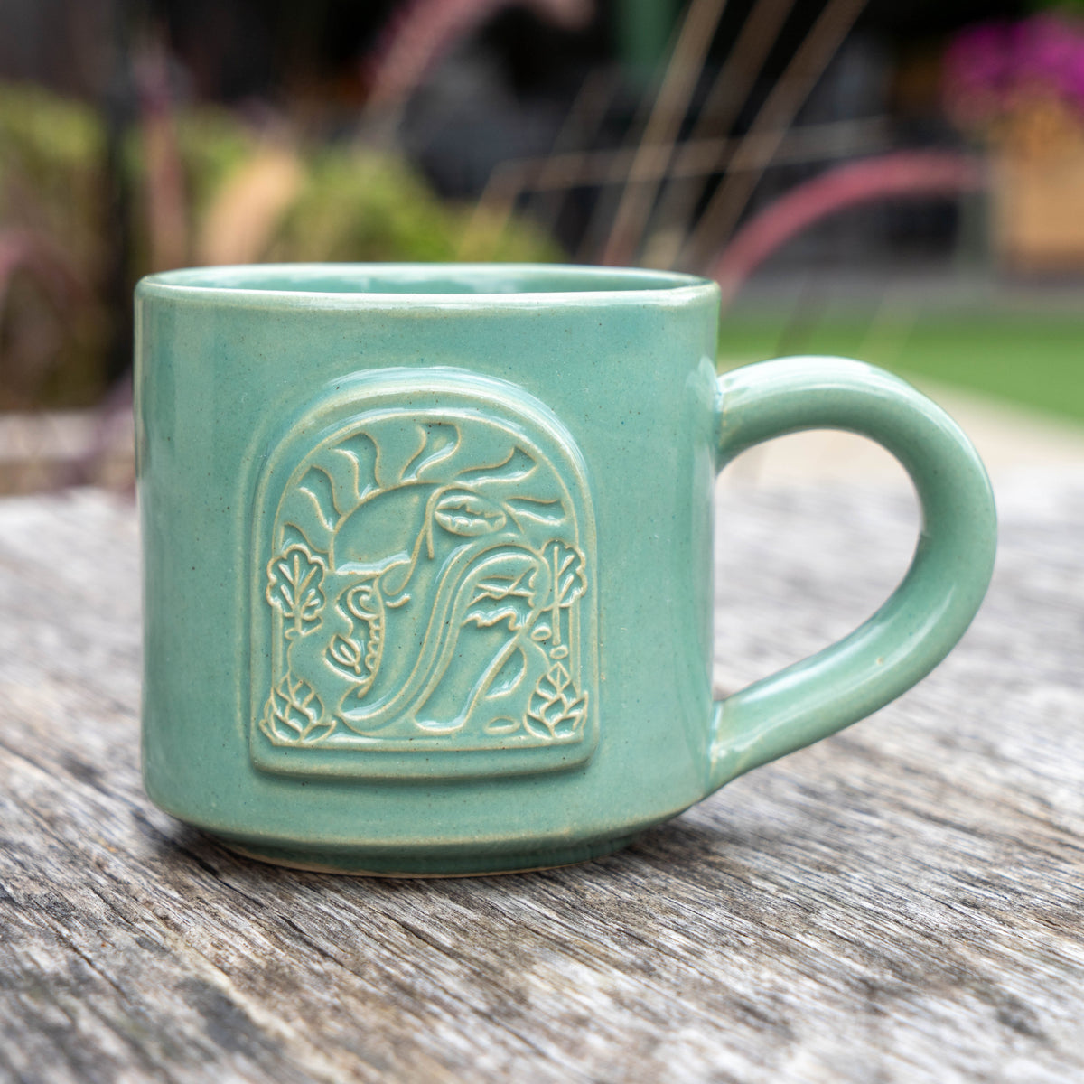 A handcrafted Dogfish Dash 12oz Ceramic Mug by Black Oak, featuring a light green finish and a raised sun and leaf design, rests on a weathered wooden surface with softly blurred greenery in the background—a perfect choice for craft beer lovers.