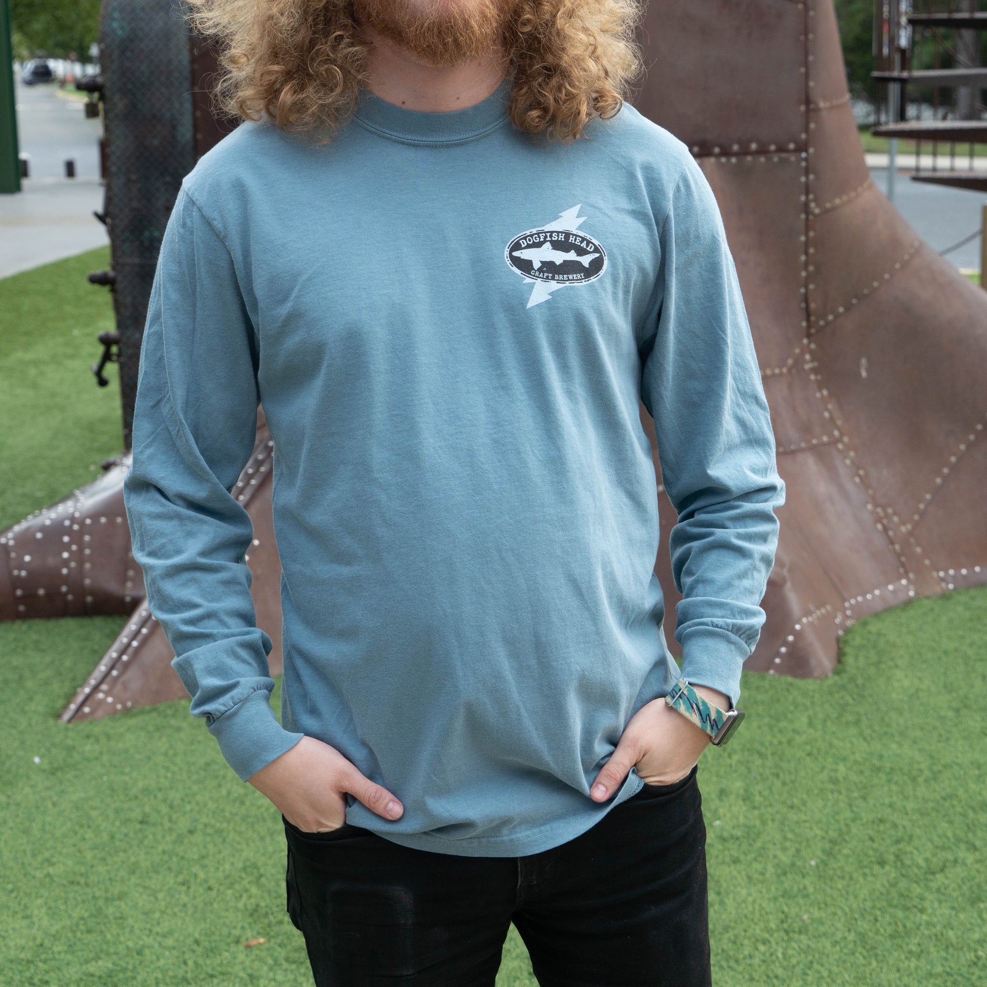 A man wearing a dusty blue long-sleeve t-shirt with a black and white shark graphic on the left chest, standing outdoors in front of a large metal sculpture on green grass.