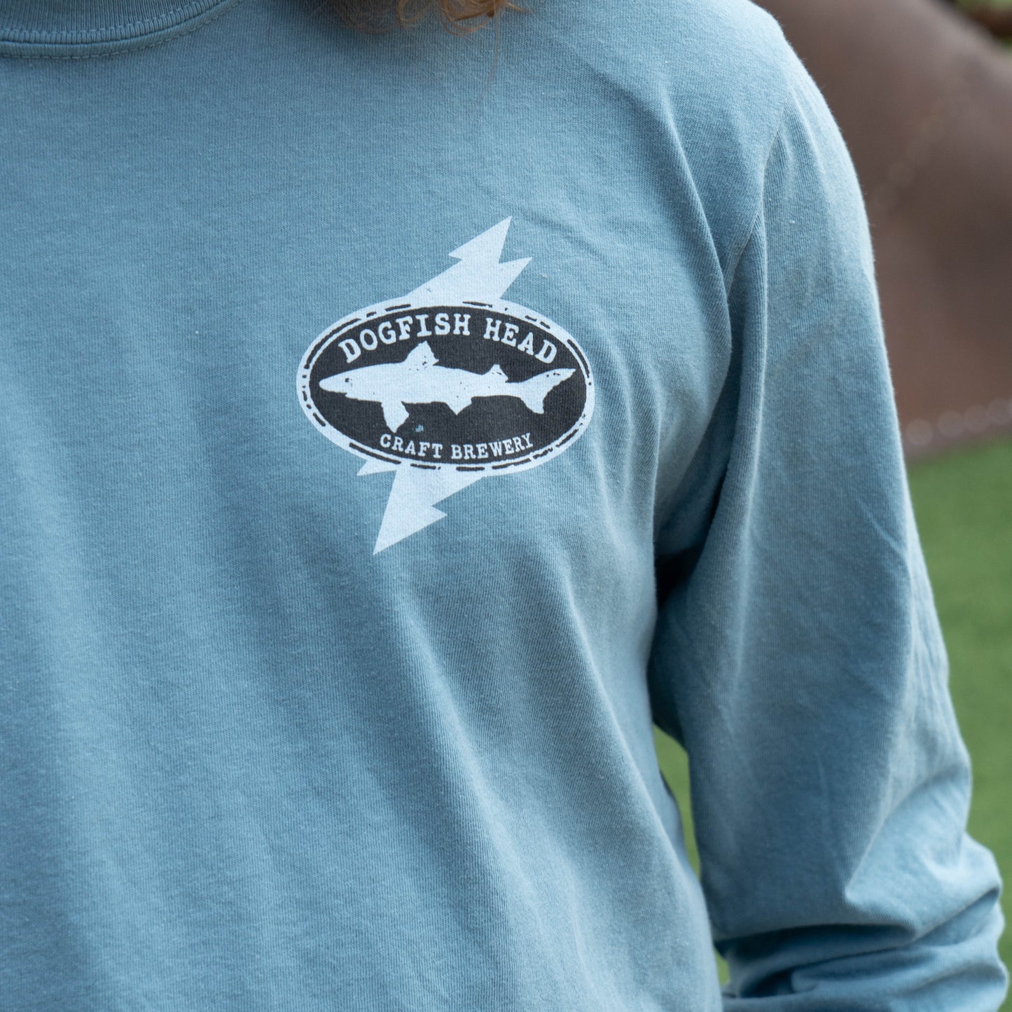 A close-up of a person wearing a heathered light blue long-sleeve t-shirt with a black and white 'Dogfish Head Craft Brewery' shark logo printed on the chest.
