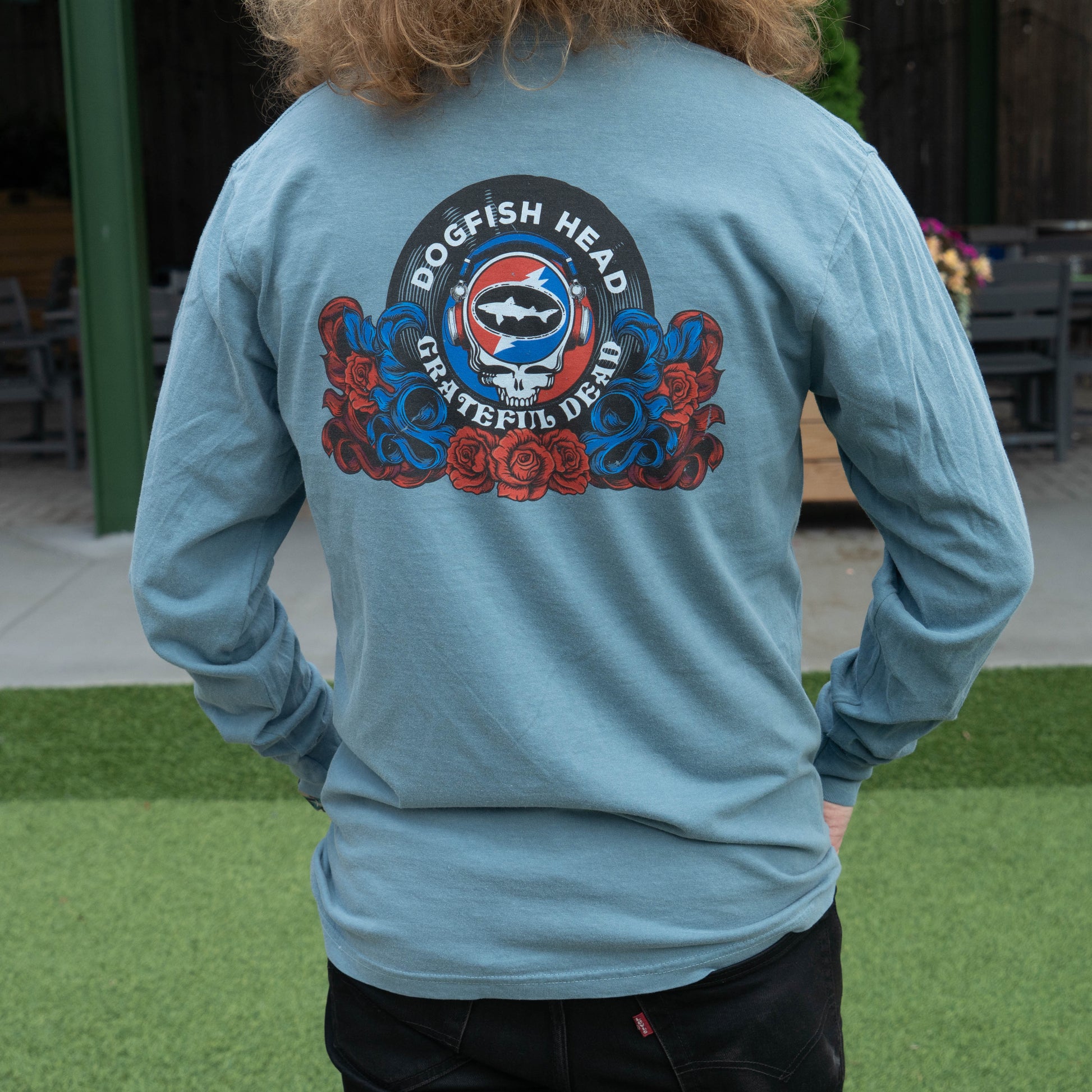 A person viewed from behind wearing a light blue long-sleeve shirt with a large circular 'Dogfish Head Grateful Dead' graphic printed on the back, featuring a skull, fish, and red and blue roses, in an outdoor patio setting.