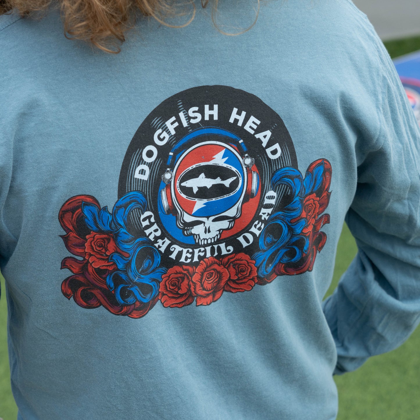 A close-up of a person wearing a light blue long-sleeve shirt, showing the back of the shirt with a large graphic print. The graphic features a circular 'Dogfish Head Grateful Dead' design with a skull, fish, stars, red and blue roses, and the words 'DOGFISH HEAD' and 'GRATEFUL DEAD'.