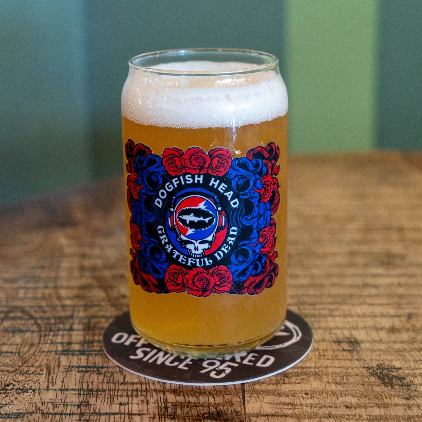 A clear pint glass filled with golden beer and topped with white foam, standing on a coaster on a wooden table. The glass has a large colorful Dogfish Head Grateful Dead label with red and blue roses and a central fish emblem.