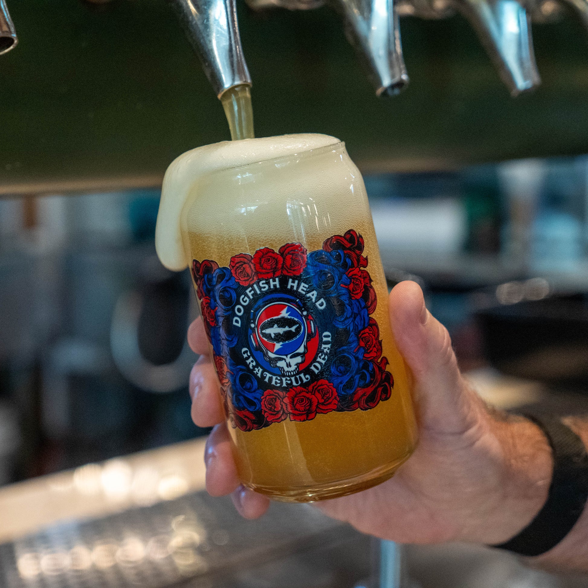 A hand holds a glass can-shaped mug filled with golden beer and a thick head of foam under a beer tap. The mug features a 'Dogfish Head Grateful Dead' label with red and blue roses and a shark logo.