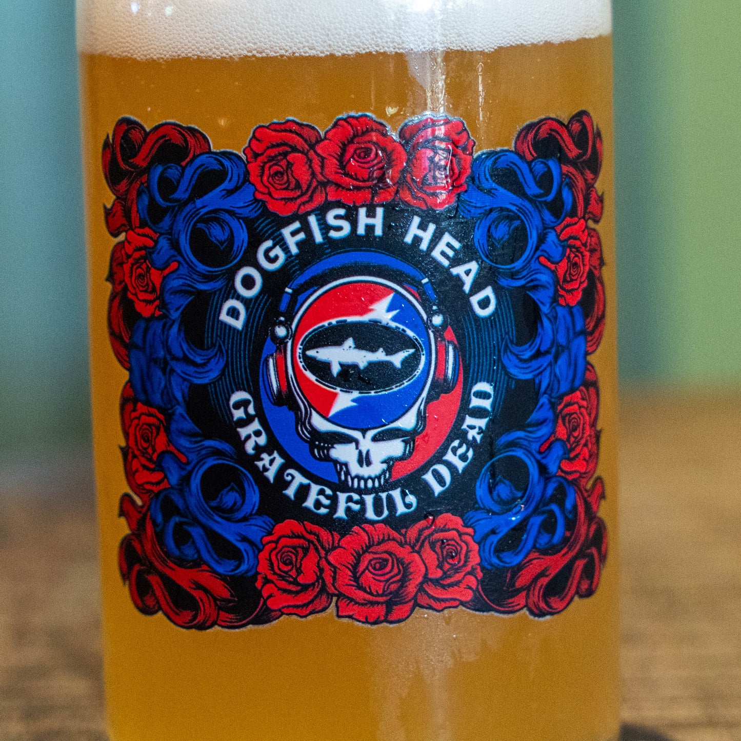 A close-up of a beer glass filled with golden beer and topped with white foam, featuring a Grateful Dead 'Dogfish Head' label with red roses and blue scrollwork.