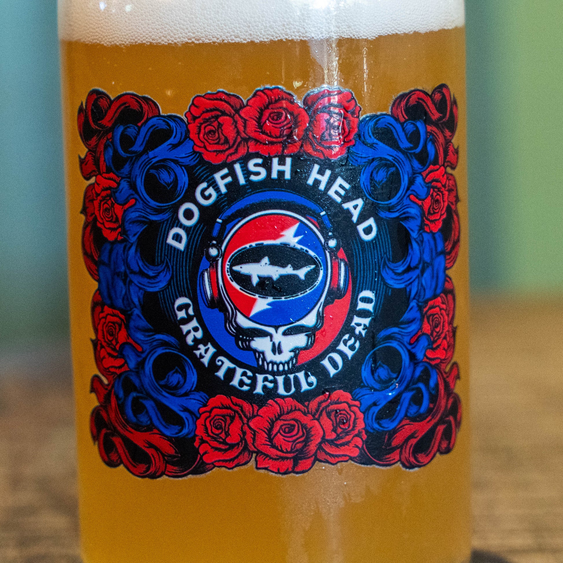 A close-up of a beer glass filled with golden beer and topped with white foam, featuring a Grateful Dead 'Dogfish Head' label with red roses and blue scrollwork.