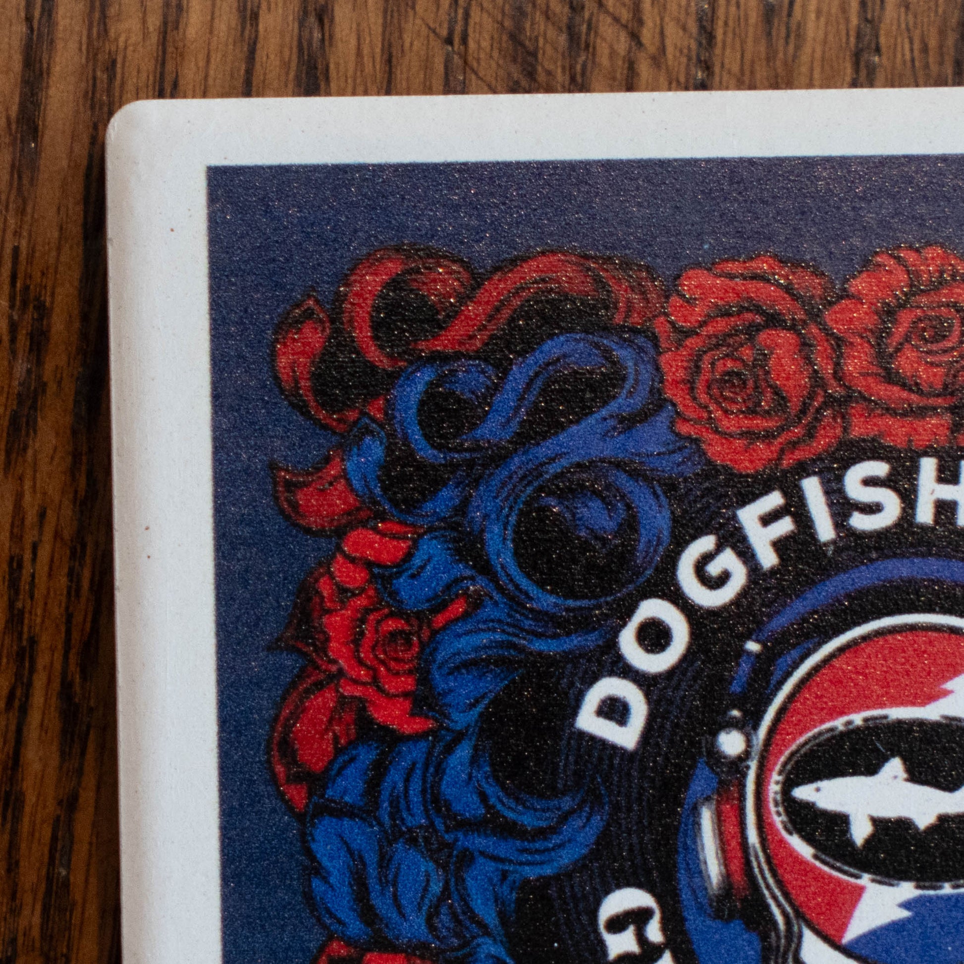 Close-up of a square coaster or tile printed with a graphic design, showing part of a Grateful Dead-style head with a fish emblem and the word "DOGFISH" in white over red and blue artwork, placed on a wooden surface.