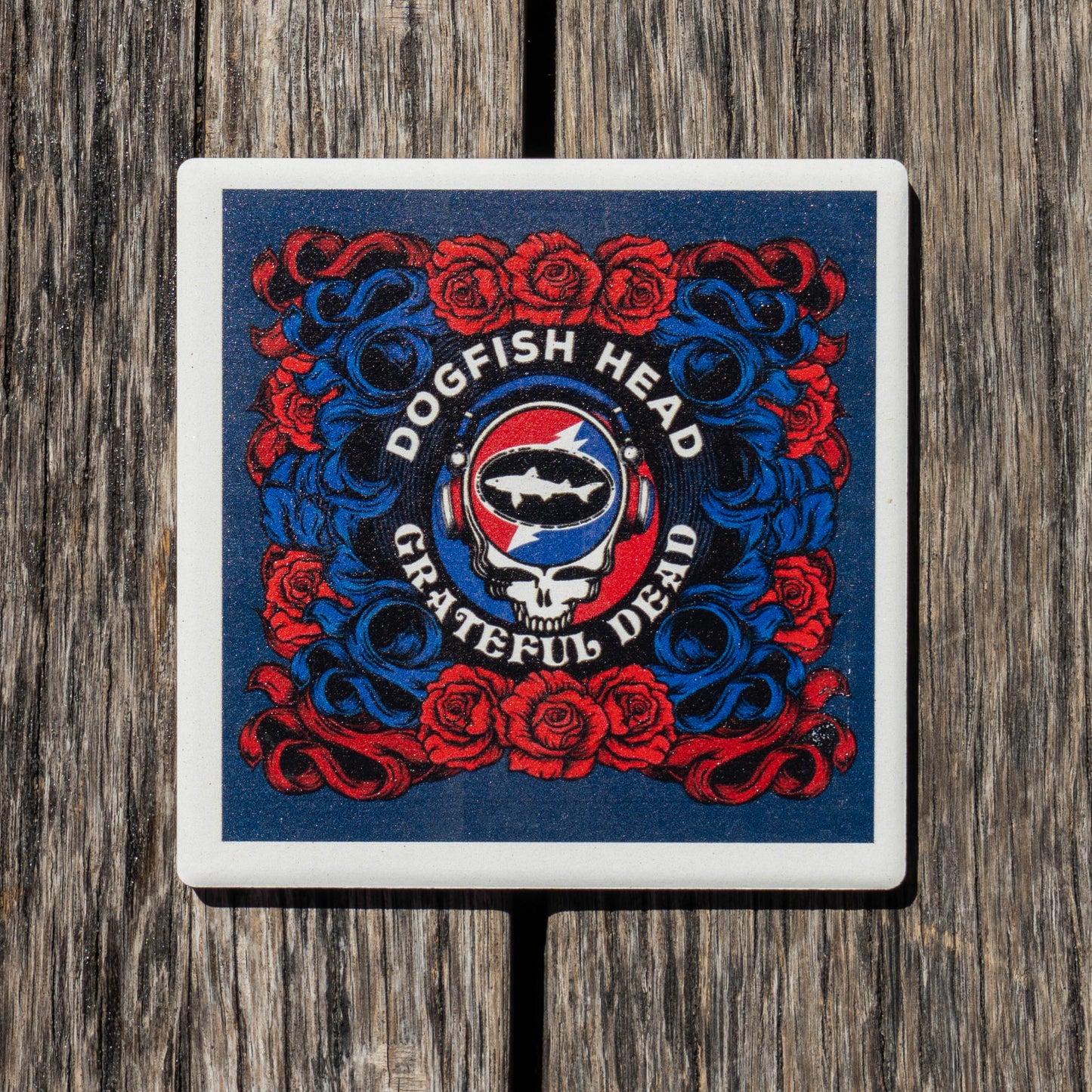A square coaster featuring the Grateful Dead Dogfish Head logo, displayed on a rustic wooden surface. The coaster has a white border and showcases the iconic red and blue Grateful Dead design with roses, a skull, and a shark.