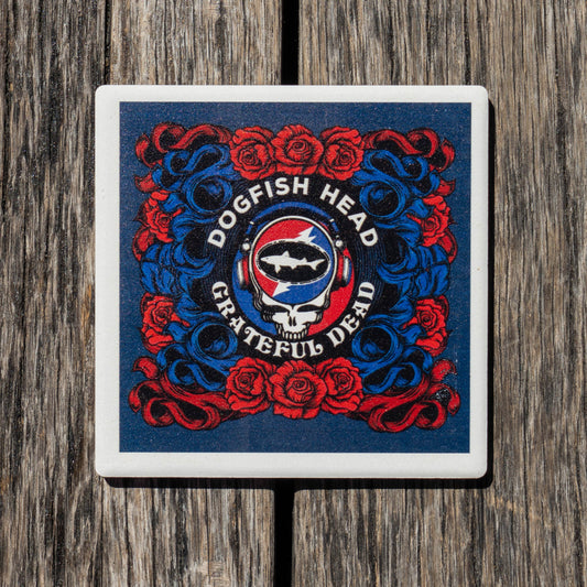 A square coaster featuring the Grateful Dead Dogfish Head logo, displayed on a rustic wooden surface. The coaster has a white border and showcases the iconic red and blue Grateful Dead design with roses, a skull, and a shark.