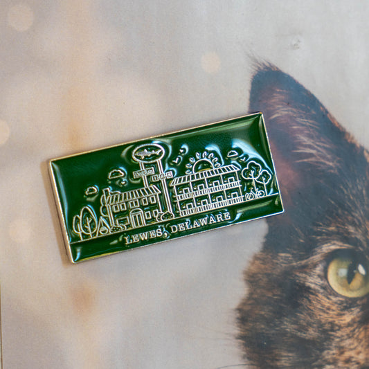 An Enamel Inn Magnet by Arrasmith Promotions LLC, featuring a green design with white building outlines and "Lewes, Delaware," sits on a surface with part of a cat’s face visible in the background.